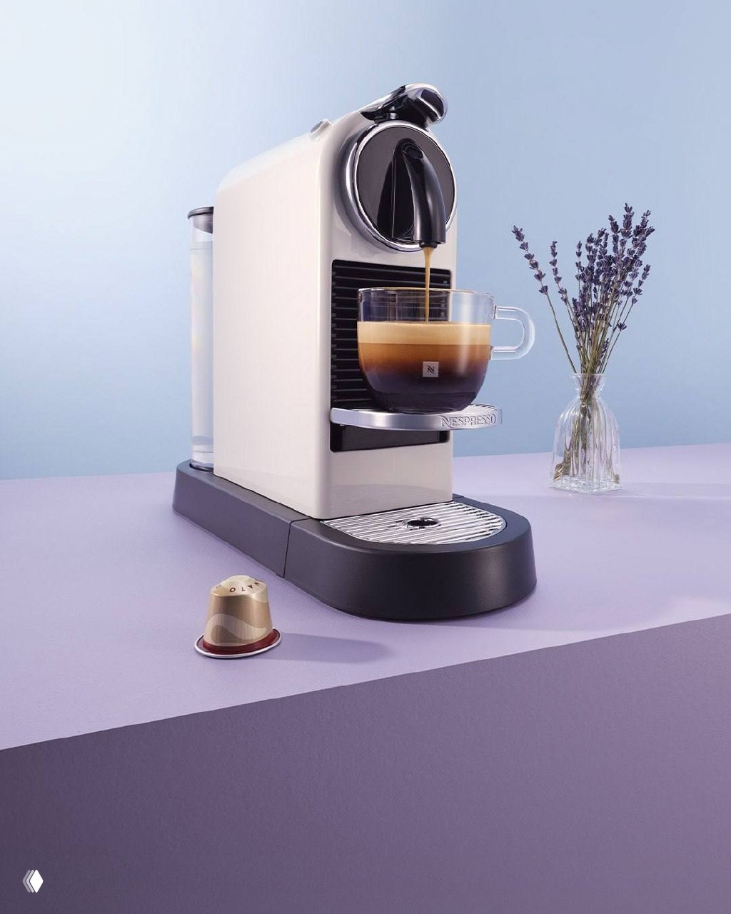Белая кофемашина Nespresso на фиолетовом фоне, рядом ваза с веточкой лаванды — рекламный кадр, где техника сочетается с цветовой концепцией кампании.