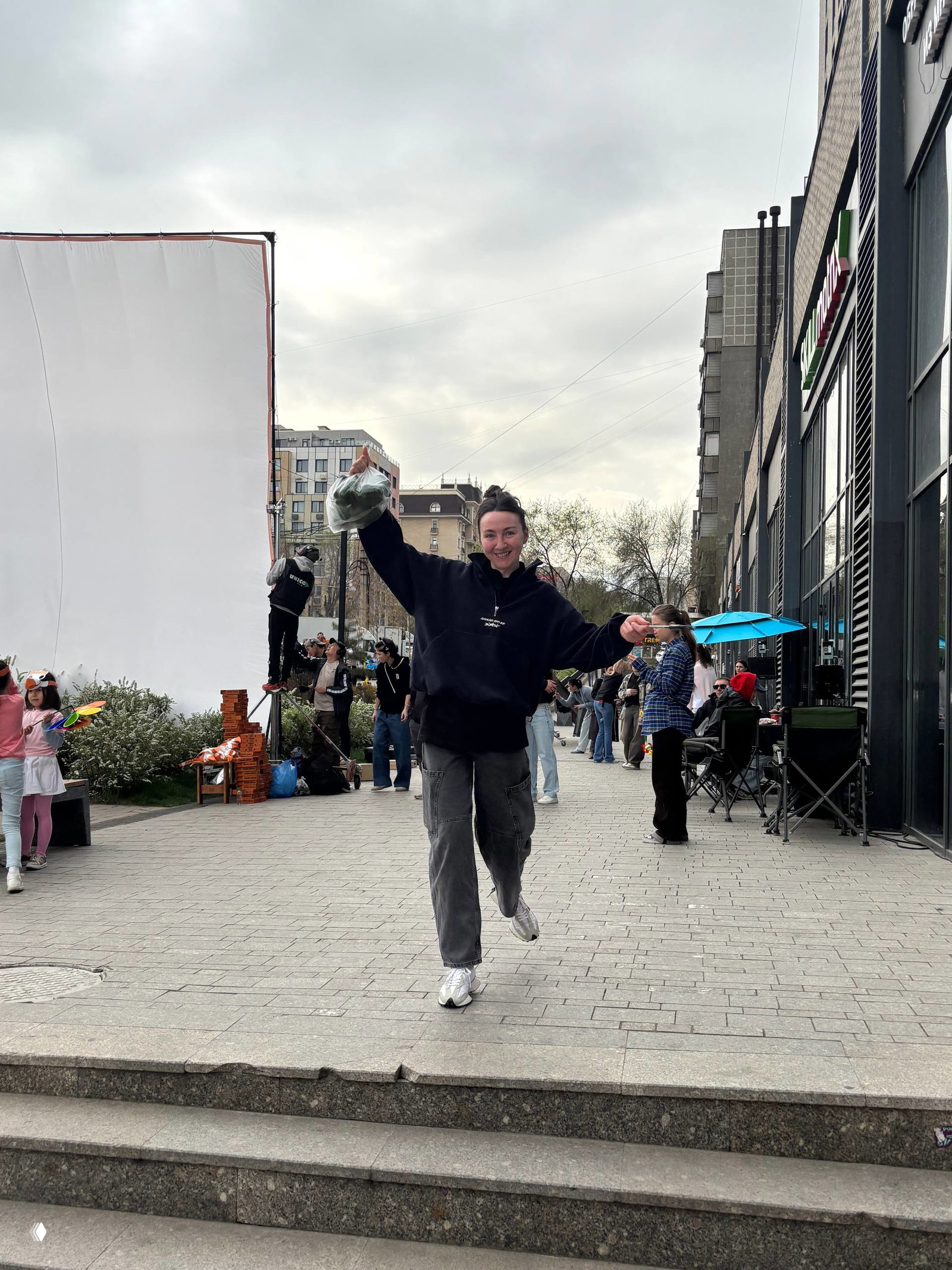 Уличный бекстейдж: бегущая участница съёмок на фоне рефлектора, ассистенты и горожане — весенняя атмосфера площадки в Алматы.