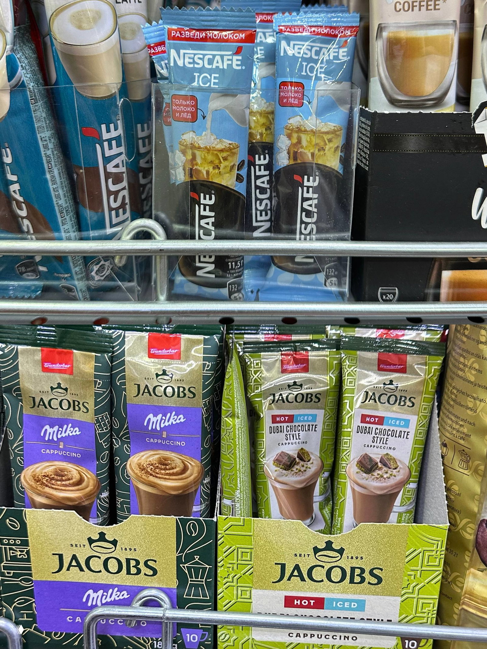 Крупный план саше Nescafe Ice и порционных упаковок Jacobs Milka/Dubai Chocolate Style на металлической полке, видны кофейные саше для горячего и холодного.