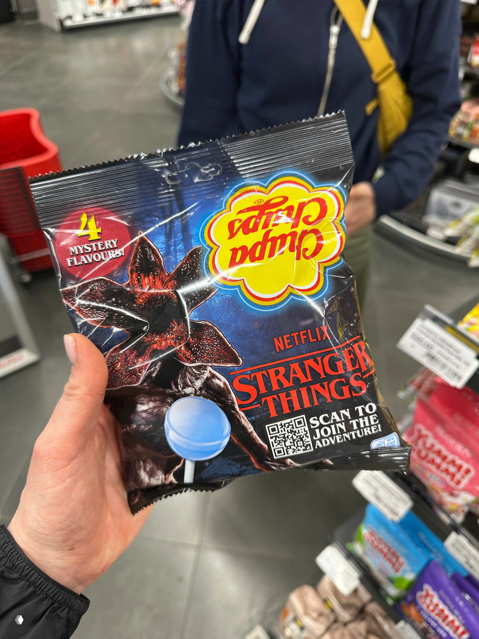 Ручной снимок пачки Chupa Chups с промо‑дизайном Stranger Things: тёмная упаковка с QR‑кодом и рекламной символикой на фоне кассовой зоны.