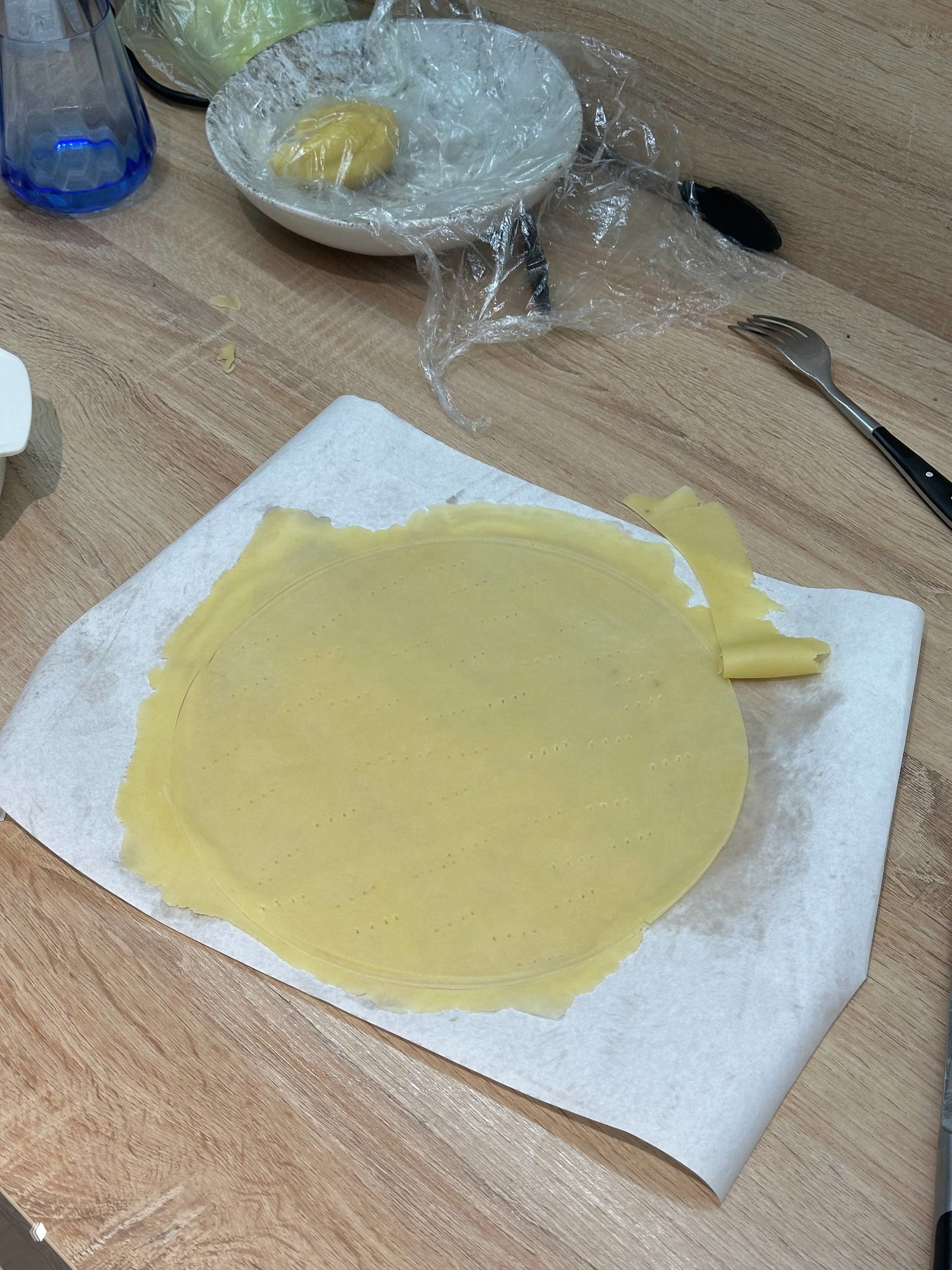 Тонко раскатанный круглый корж теста на пекарской бумаге, вид сверху; на рабочем столе также миска с оставшимся тестом и кухонные принадлежности.
