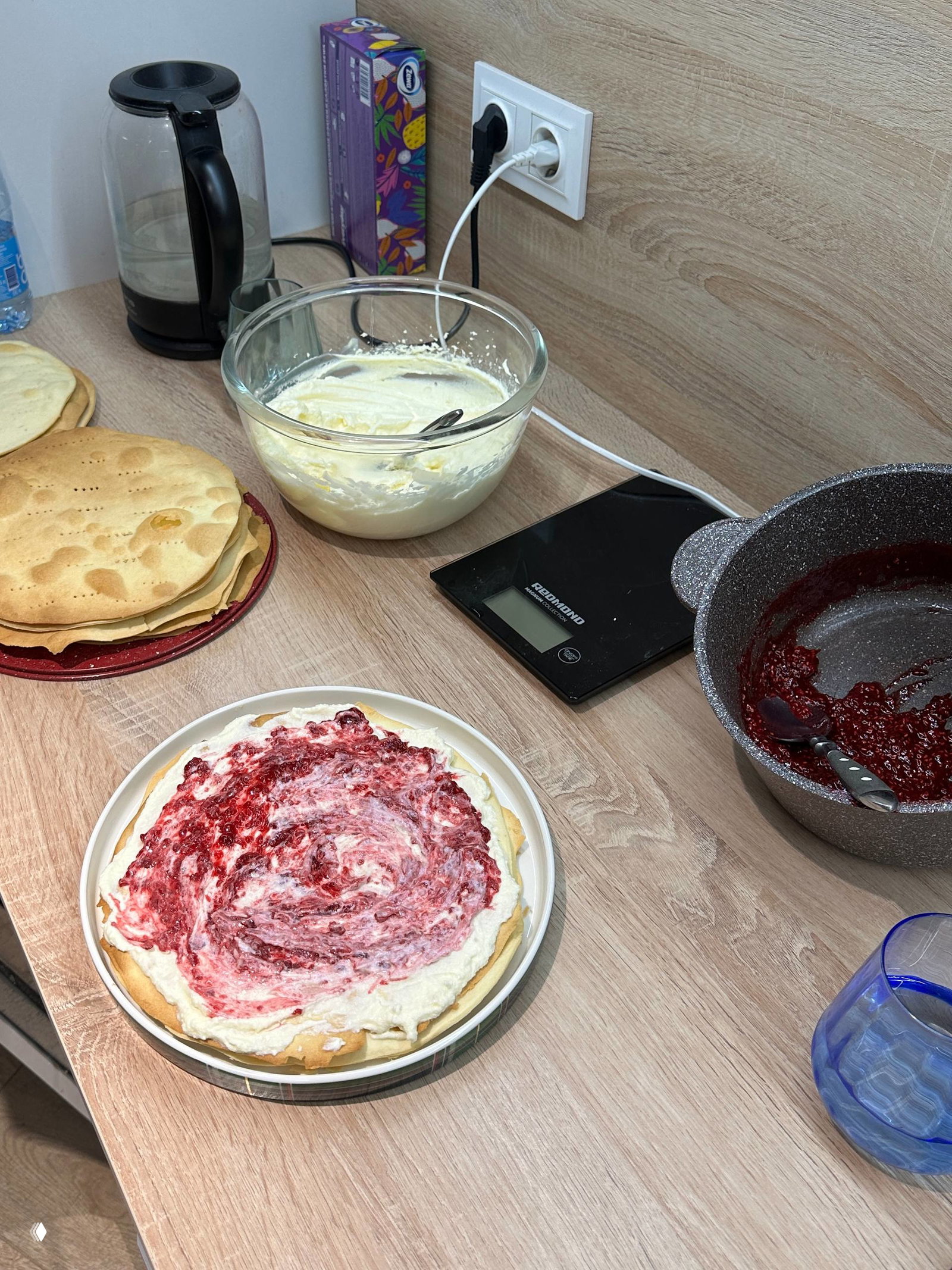Рабочий стол с готовыми тонкими коржами, миской с кремом и сотейником с малиновым соусом; этап сборки торта и распределение начинки.