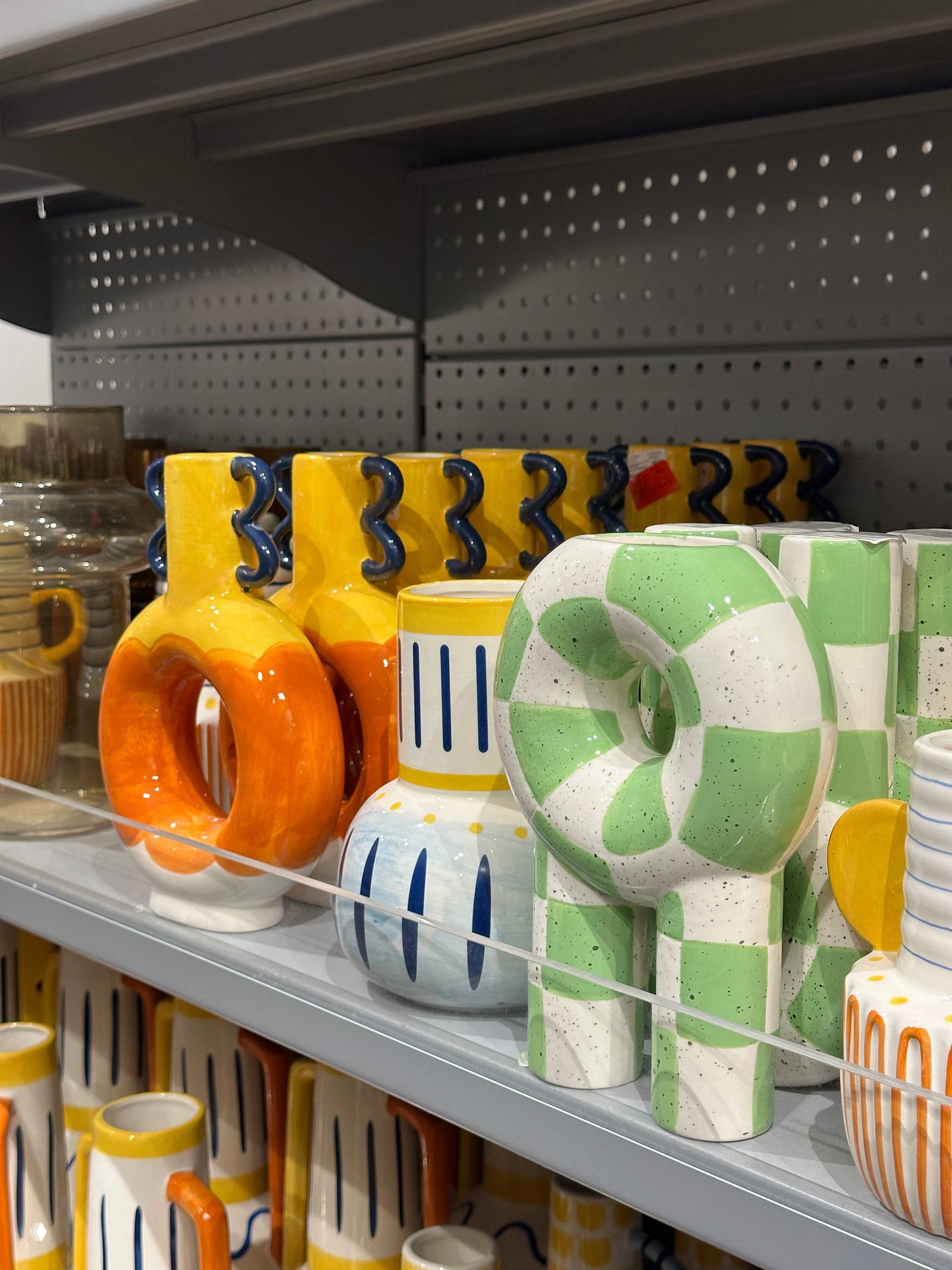 Ряд ярких декоративных ваз и керамических сосудов — оранжевые, жёлтые и зелёные предметы с причудливыми ручками, расставленные на торговой полке.