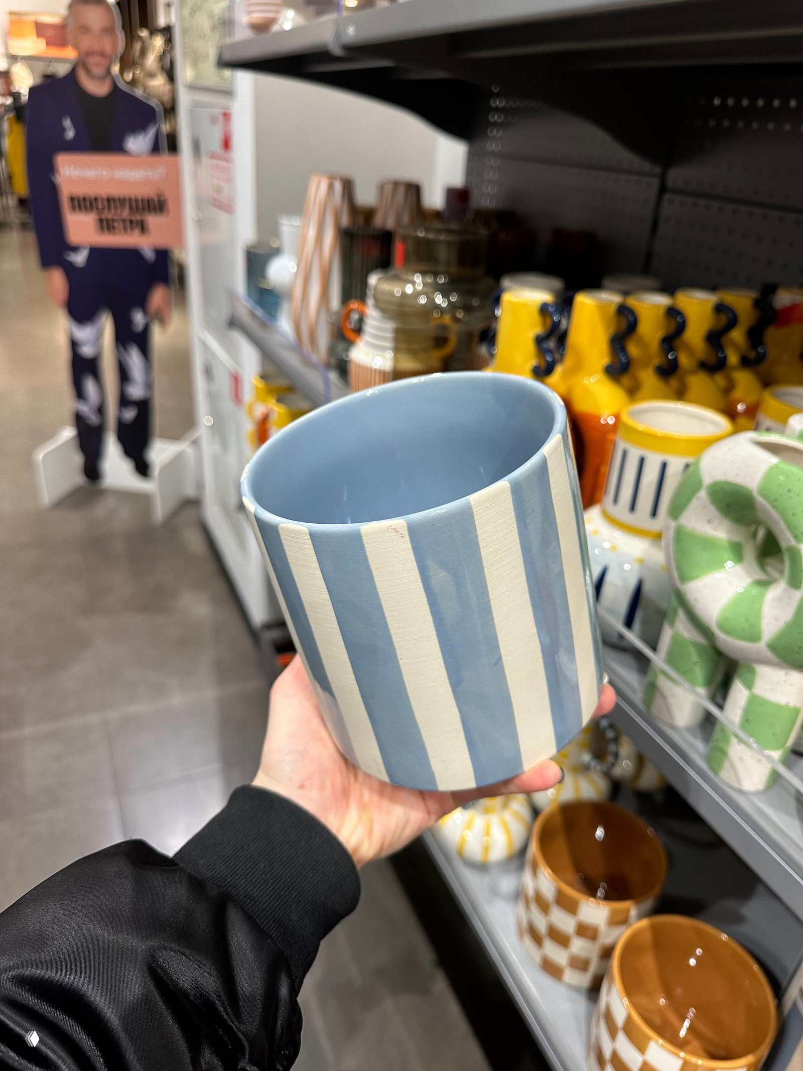 Крупный план полосатого керамического стакана в руке автора на фоне полок с разноцветной посудой и витрины магазина Offprice.