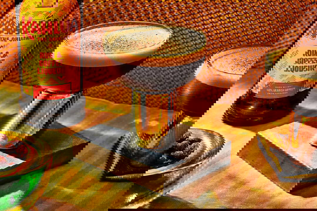 Крупный план высоких бокалов с густым тёмным «кофе», бутылка Kahlúa на заднем плане и золотистые подставки — фуд‑фотосъёмка для рекламы напитка.