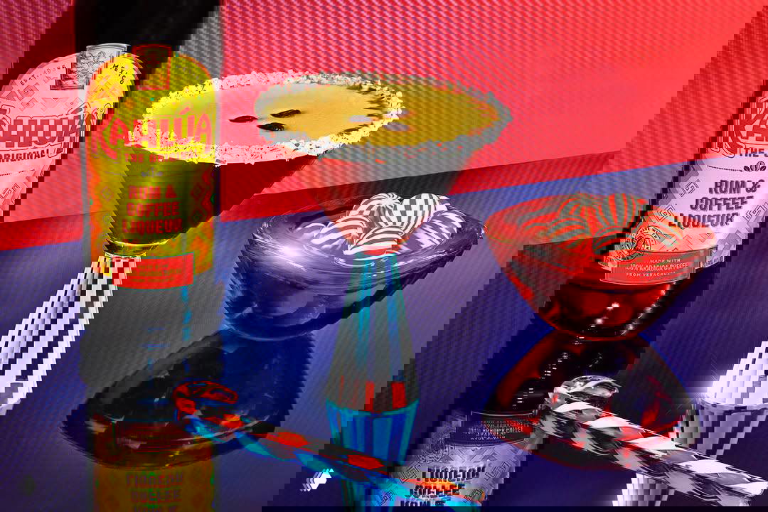 Коктейль в рюмке с густым кофейным слоем и бутылка Kahlúa рядом на цветном фоне; акцент на текстуре и естественном внешнем виде напитка.