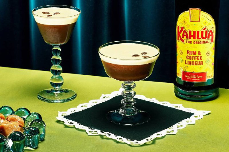 Два низких бокала с густым кофейным напитком, лёгкая пена и капли украшения рядом с бутылкой Kahlúa на зелёной поверхности — съёмочная стилизация.