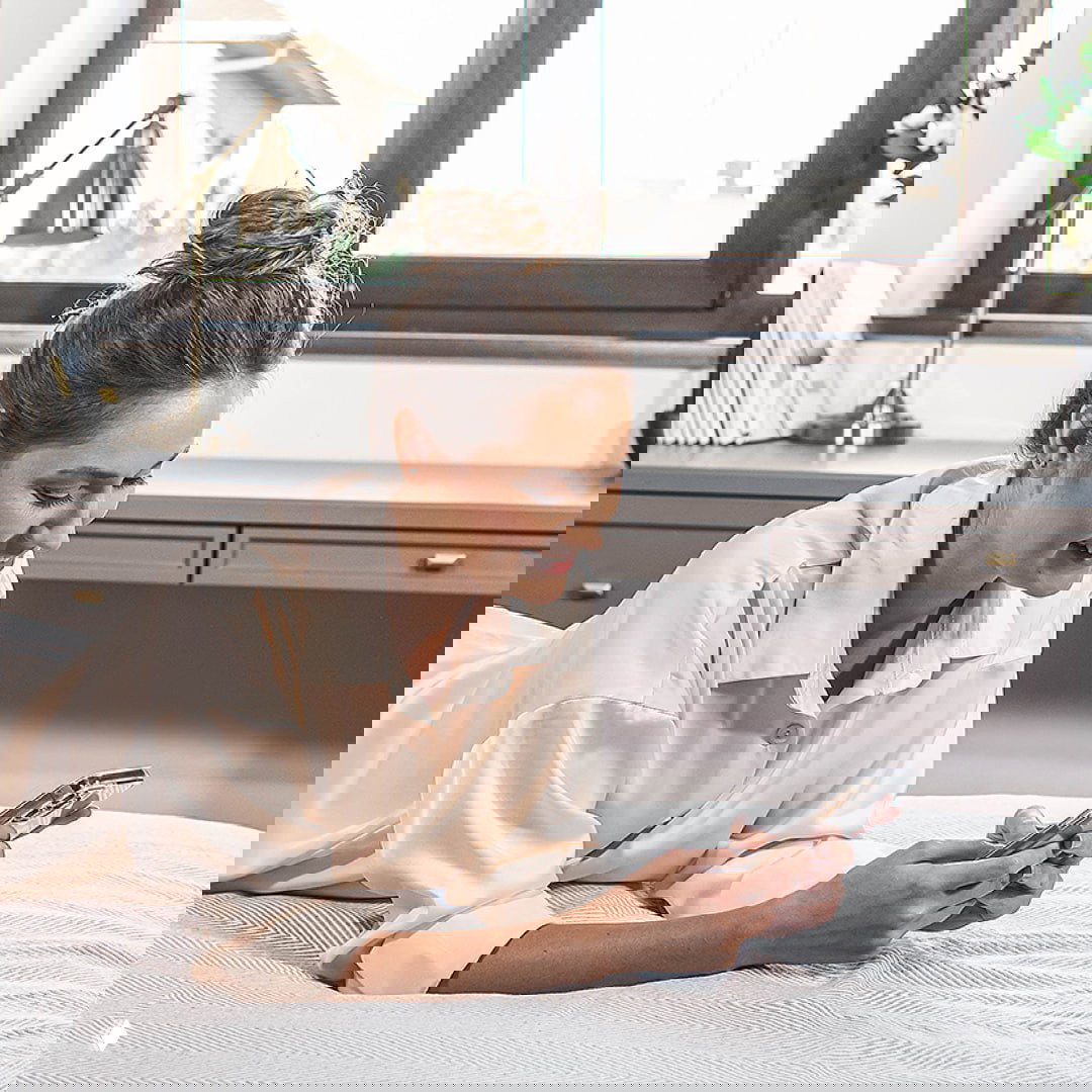 Женщина лежит на кровати и смотрит в смартфон, символизируя удобство мобильного шопинга и выбора продукции РЕХАУ через телефон.