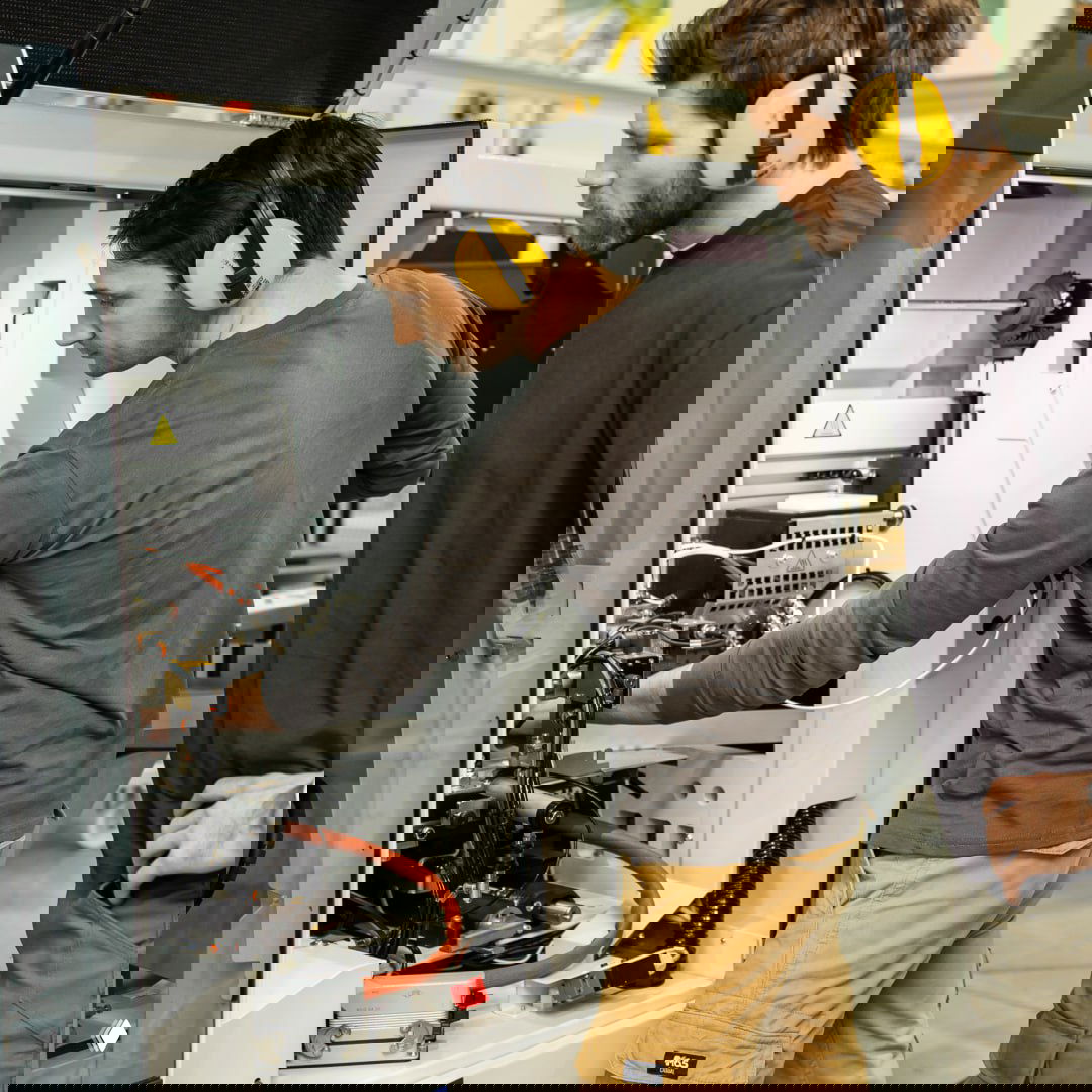 Два рабочих в цеху осматривают внутреннюю часть станка, используют защитные наушники и инструменты при проверке, обсуждая наладку и диагностику оборудования.