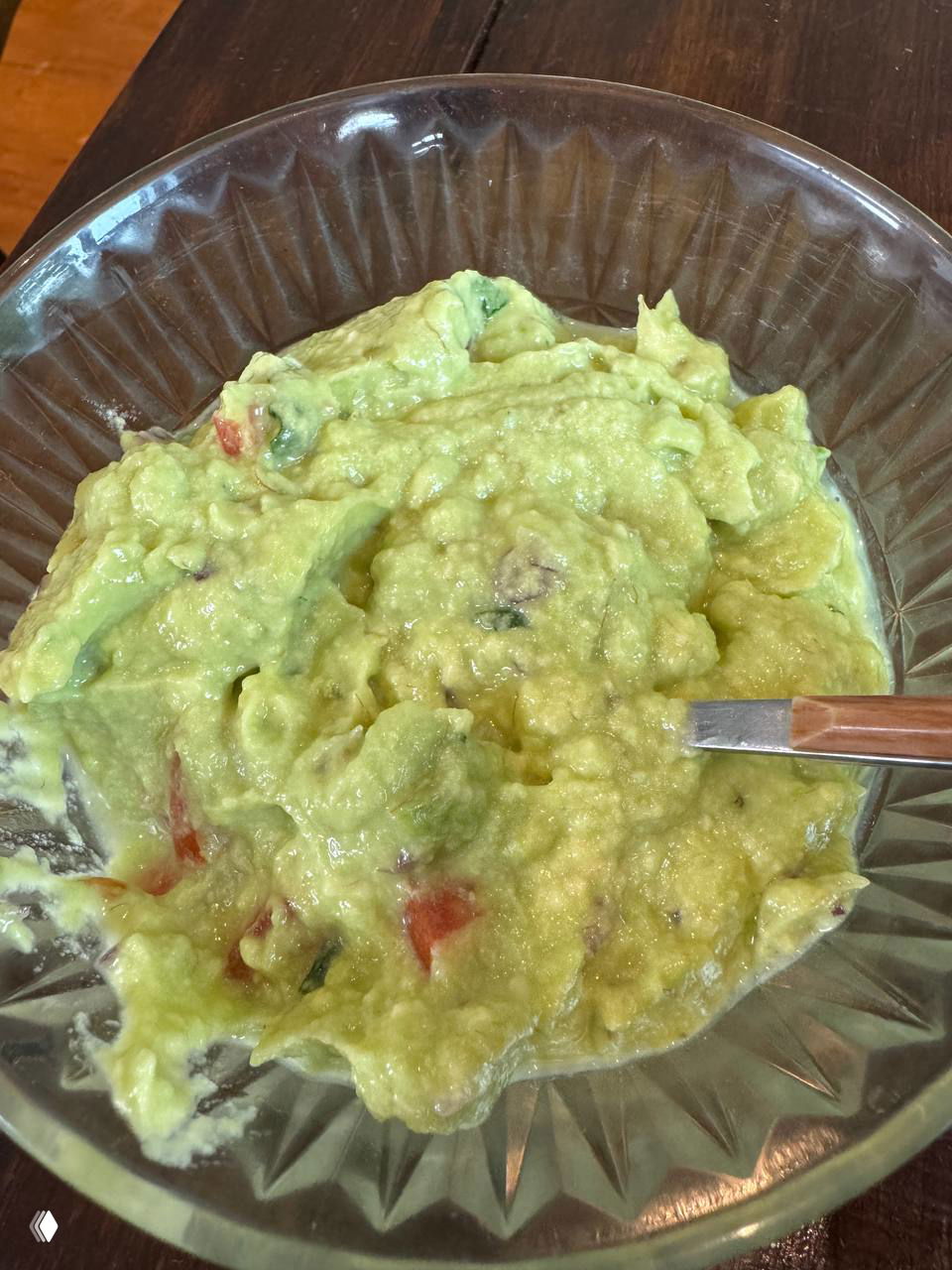 Стеклянная миска с гуакамоле светло-зеленого цвета, водянистой текстуры, видны кусочки помидора и ложка — домашнее пюре из авокадо.