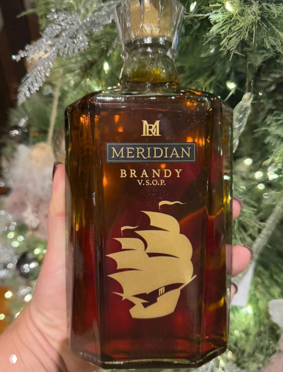 Бутылка бренди Meridian V.S.O.P. с золотым изображением парусного корабля на этикетке, держат на фоне праздничной ёлки и декора.