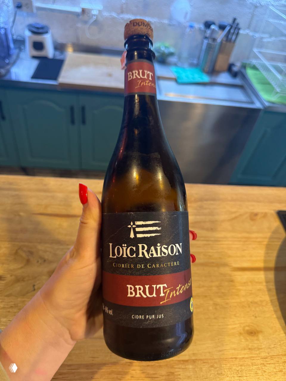 Бутылка французского сидра Loïc Raison Brut в руке: этикетка и пробка видны на фоне барной стойки и кухни.