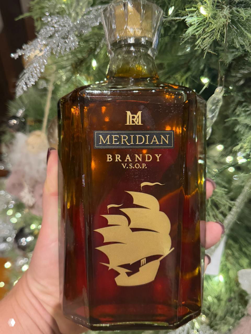 Крупный план бутылки Meridian Brandy V.S.O.P. с золотистым изображением корабля на этикетке, бутылка сфотографирована на фоне ёлки и огней.