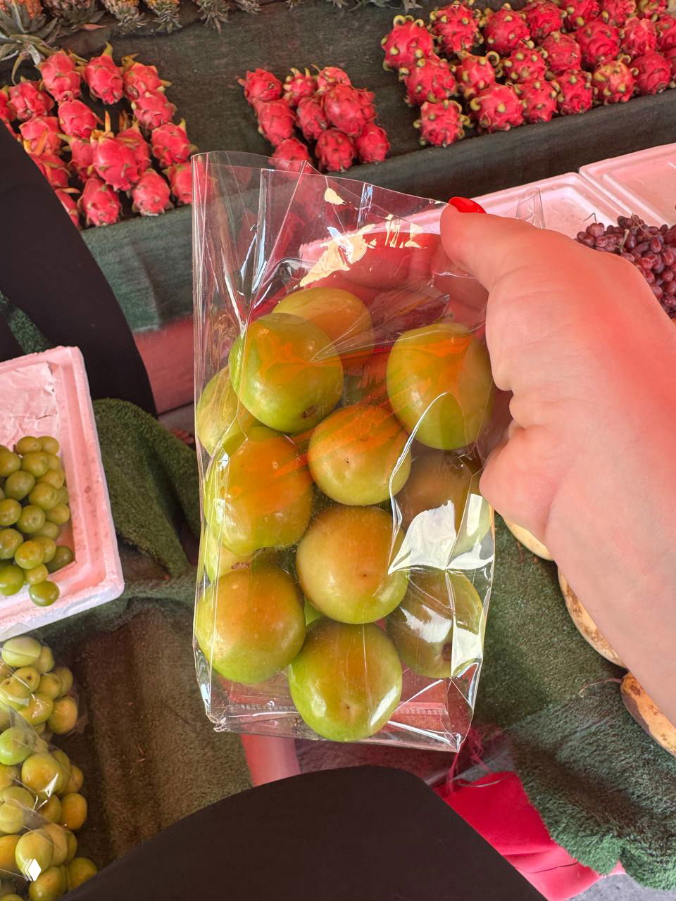 Пакет с зелёными плодами путса (зизифус) в руках продавца на рынке Самуи; на заднем плане драгонфруты и прилавок.