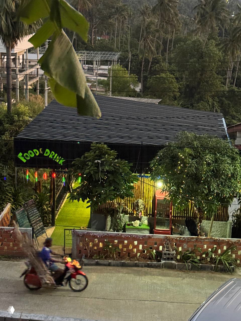 Ночная фасадная часть новой кафешки на Ламай со светящимися гирляндами и вывеской «Food & Drink», мотоциклист проезжает по дороге внизу.
