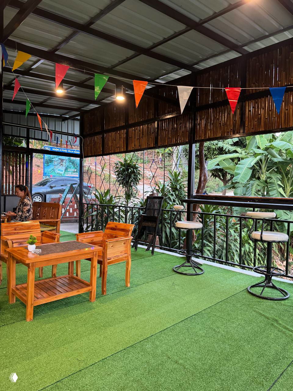 Уютная открытая терраса кафе с деревянной мебелью, искусственной травой, высокими стульями и растениями — пространство для семейных встреч и закатов.