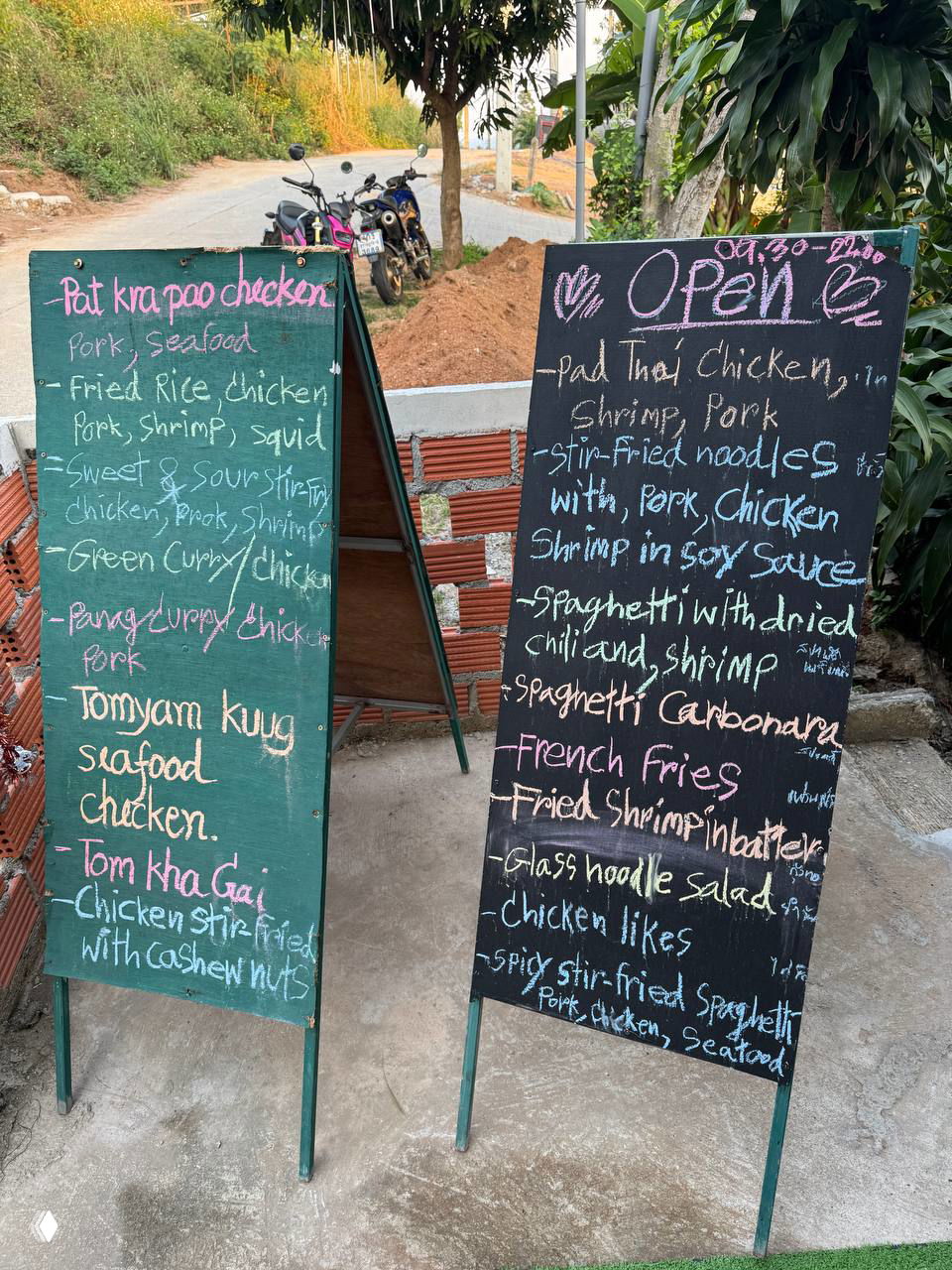 Две меловых доски-меню у входа в кафе с перечнем блюд на английском, разнообразные позиции и фрагмент улицы рядом с припаркованными мотоциклами.