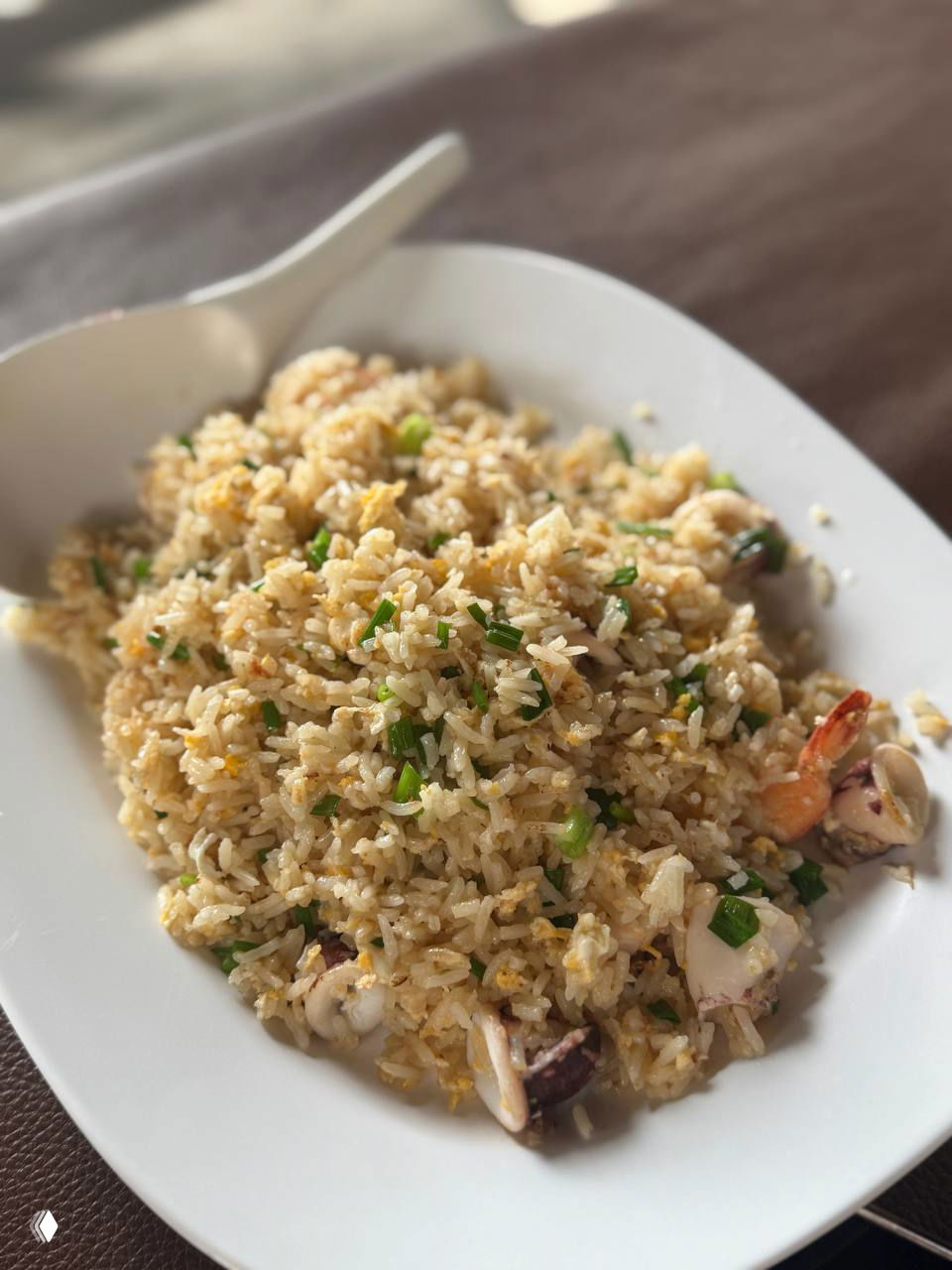 Большая порция жареного риса с морепродуктами (креветки, кальмар), вид крупным планом на белой тарелке — зерна, зелёный лук и сочные кусочки.
