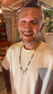 Улыбающийся молодой мужчина в светлой рубашке внутри ресторана — портрет хозяина в тёплой атмосфере с интерьером и огнями на заднем плане.