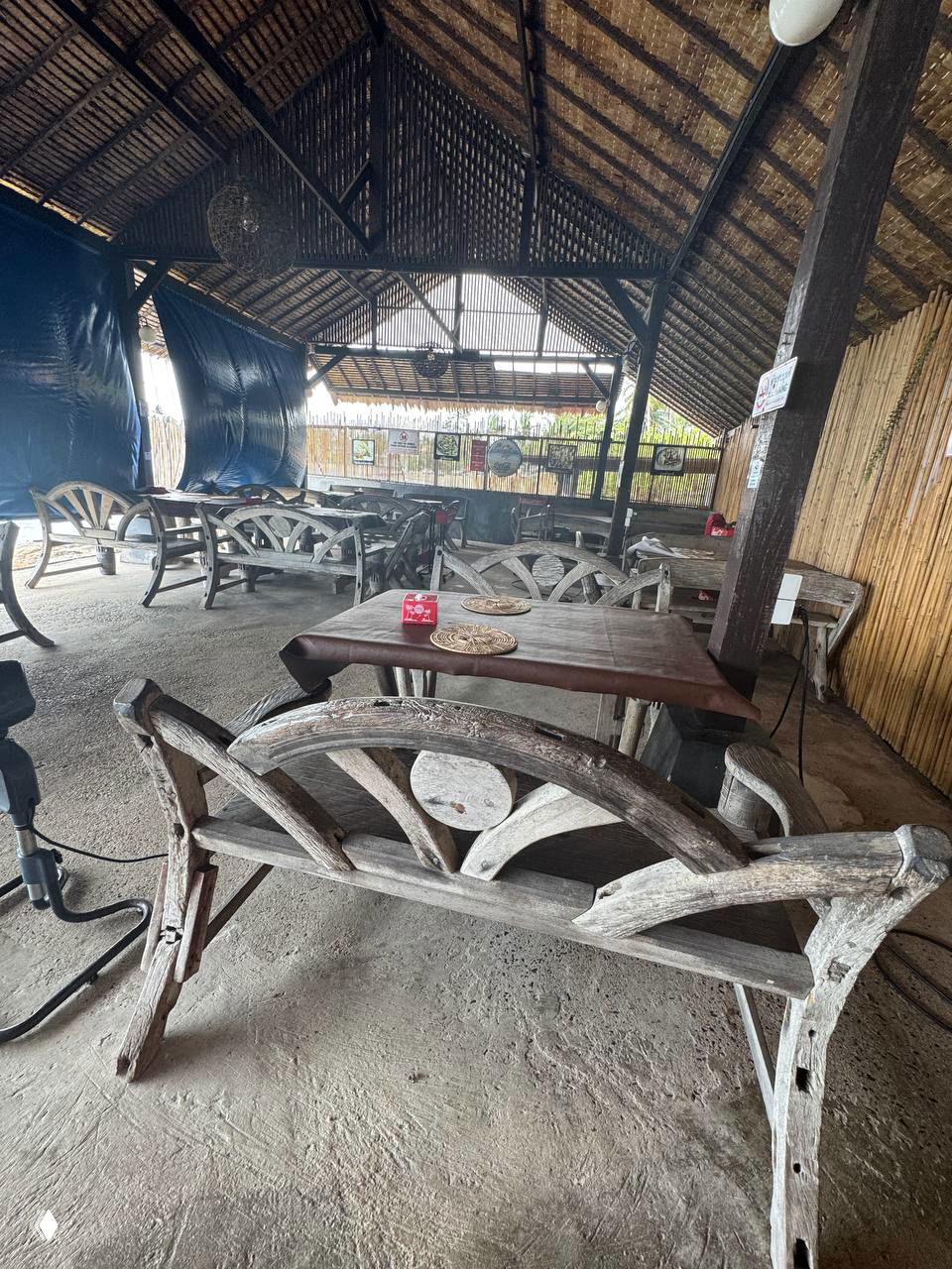 Внутреннее пространство ресторана: пустые деревянные лавки и столы под соломенной крышей, атмосфера простоты и местного уюта.
