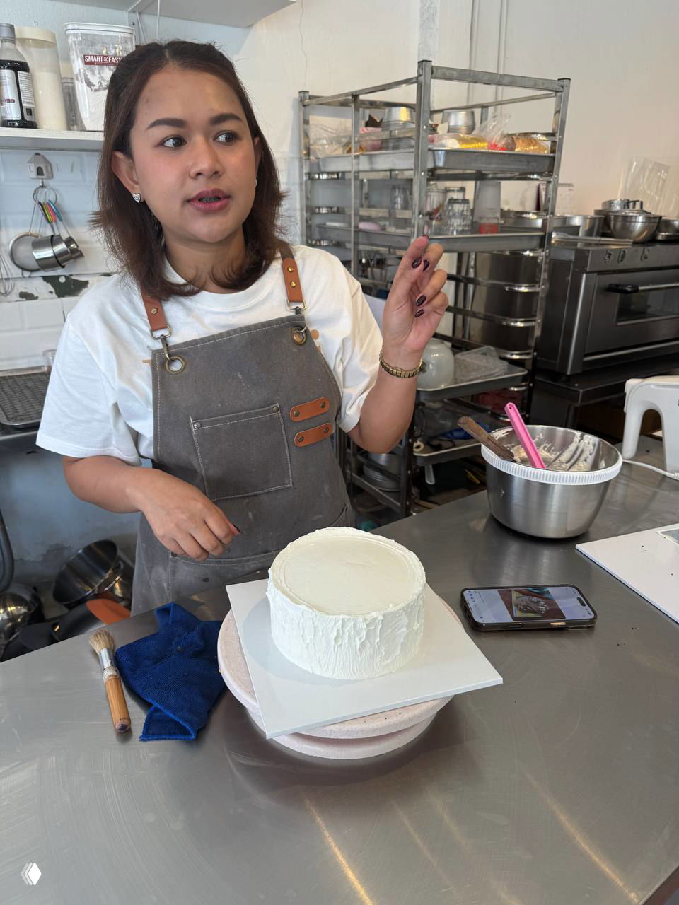 Юи в фартуке у стойки кондитерской, демонстрирует торт на поворотной подставке в рабочей зоне с инструментами и мисками.