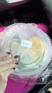 Круглый пластиковый контейнер с готовым ланчем (карри со свининой), вид сверху; рука с тёмным маникюром держит упаковку на фоне салона машины.