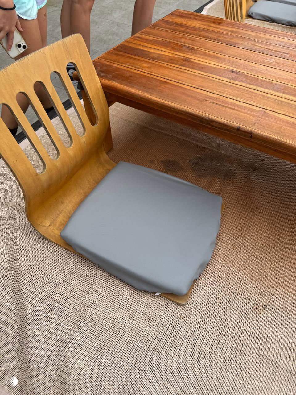 Низкий деревянный стул с серой подушкой у деревянного стола на веранде кафе Highland Paradise; рядом видны ноги посетителей и полыеранда.