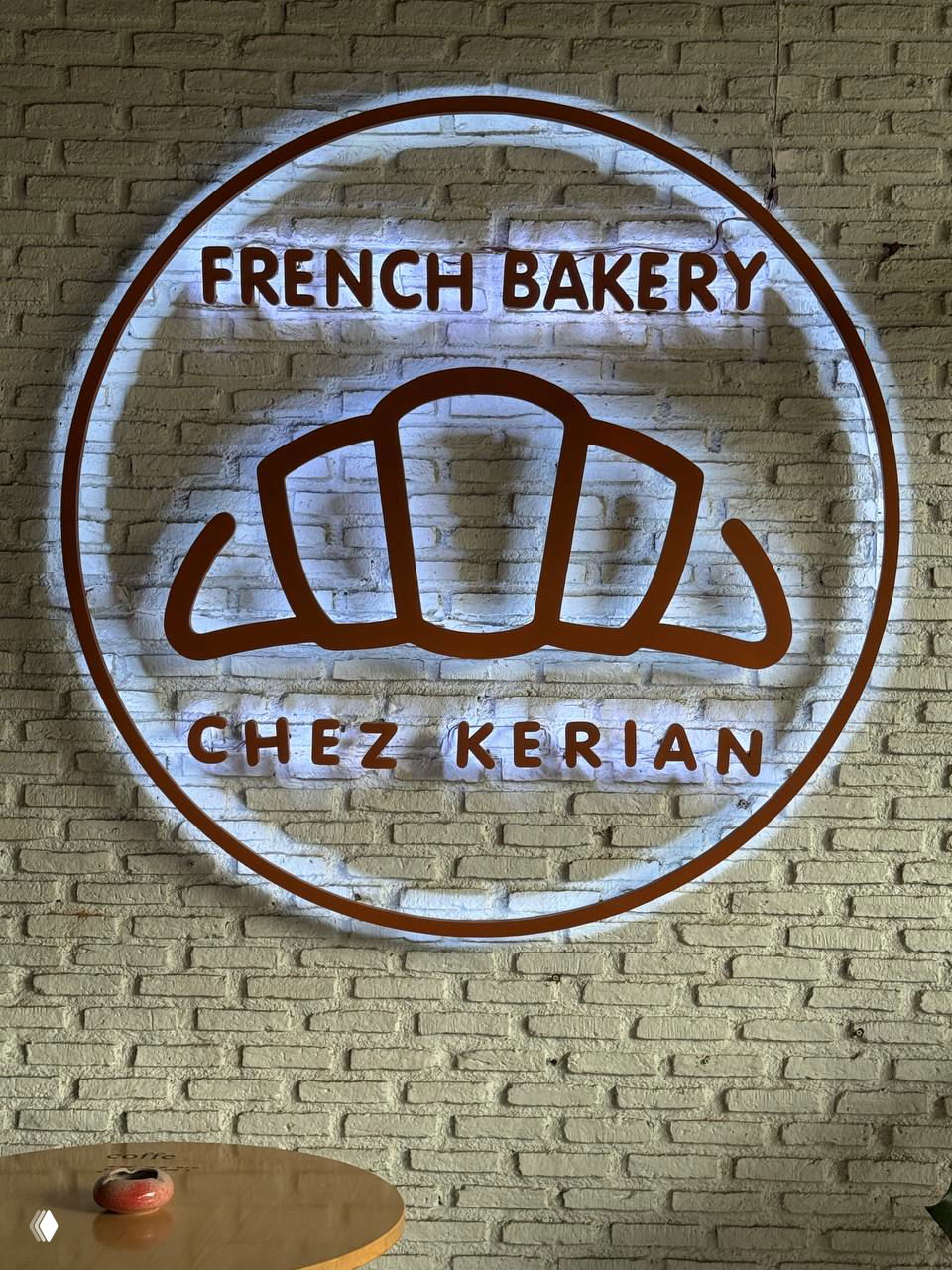 Неоновая вывеска на кирпичной стене с логотипом круассана и надписью French Bakery — Chez Kerian, интерьер у стола.