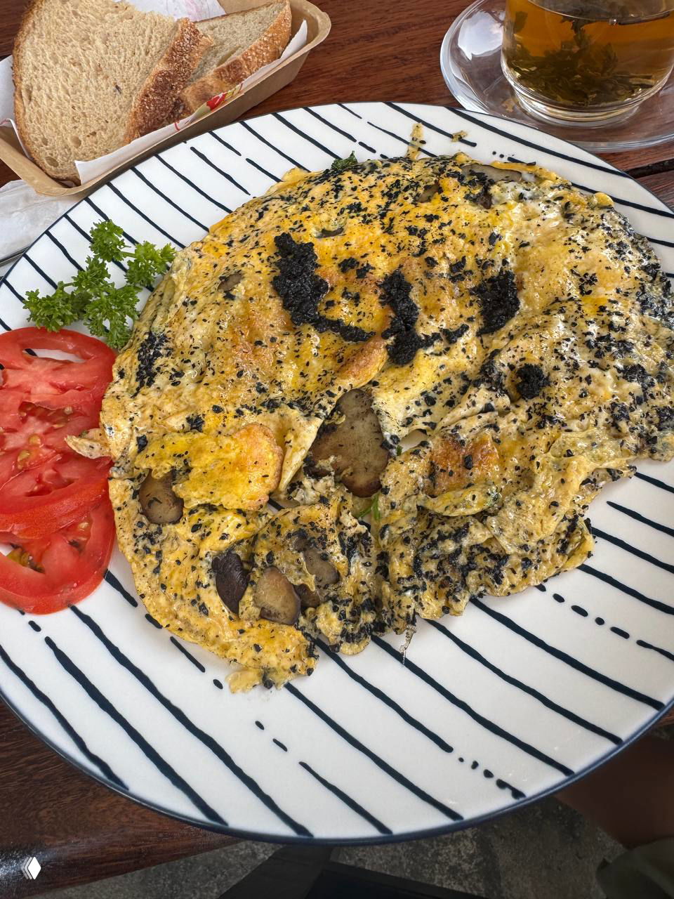 Омлет с трюфельной пастой на белой тарелке в чёрную полоску; рядом ломтики помидора и хлеб — блюдо выглядит простым, с чёрными вкраплениями трюфельной пасты.