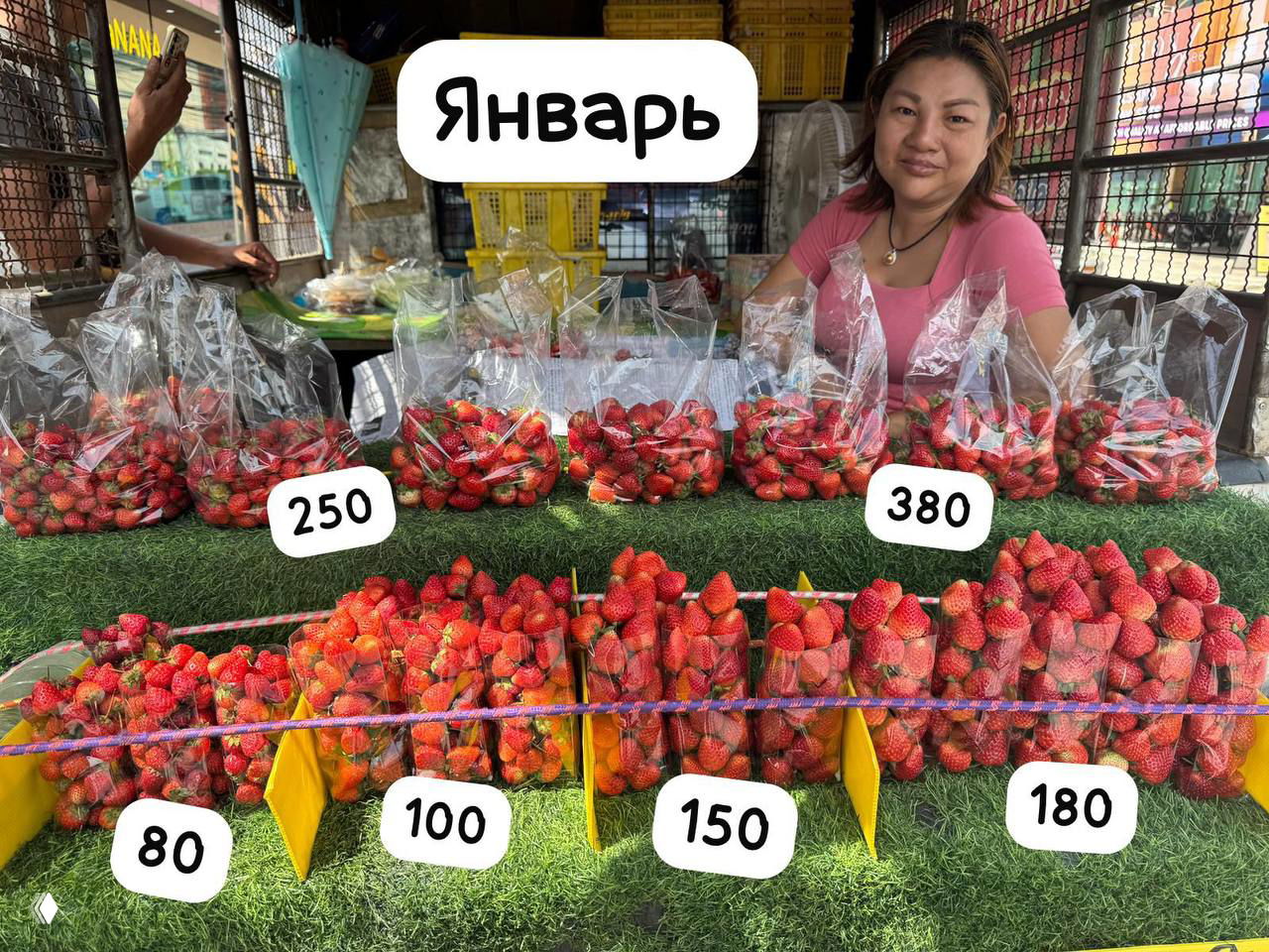 Продавщица и прилавок с клубникой (январь): прозрачные пакеты с ягодами и видимые ценники 80, 100, 150, 180 и 380 бат.