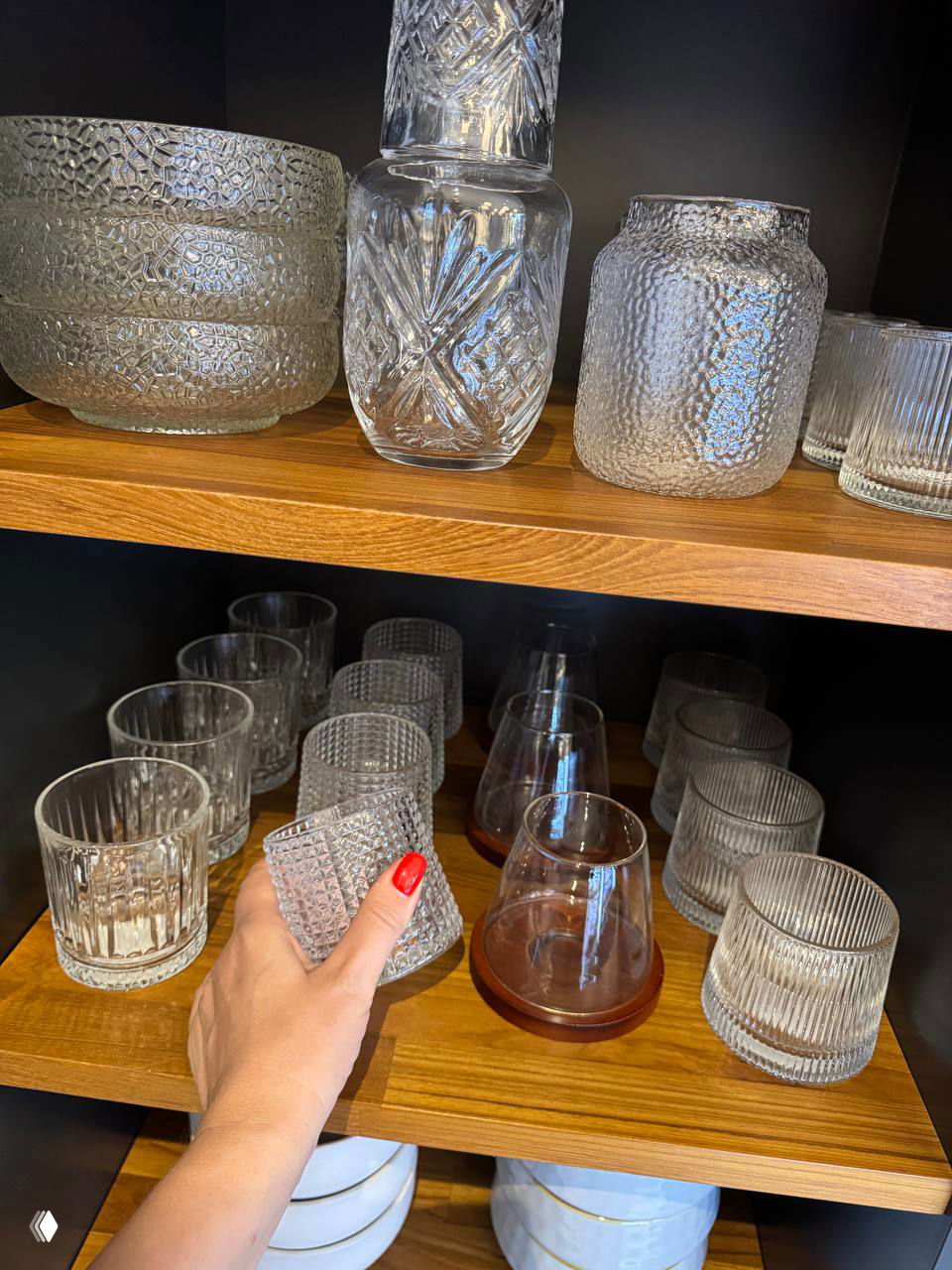Крупный план стеклянных стаканов и бокалов на полке; в кадре рука с красным маникюром держит один стакан, акцент на текстуре стекла.