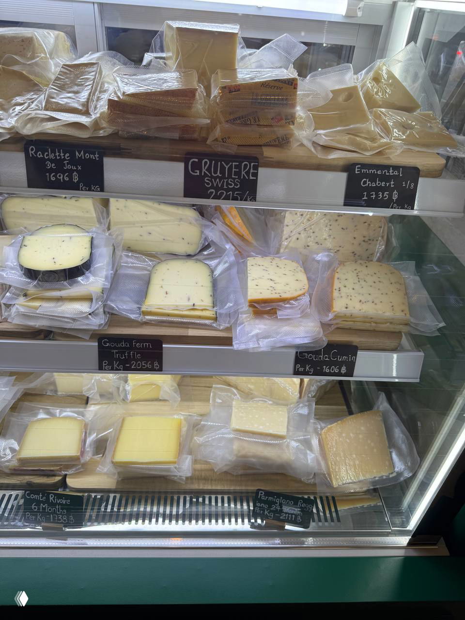 Витрина с кусками французских сыров в вакуумной упаковке: разные сорта (грюер, гауда и др.) с ценниками в батах на фоне стеклянной стойки.
