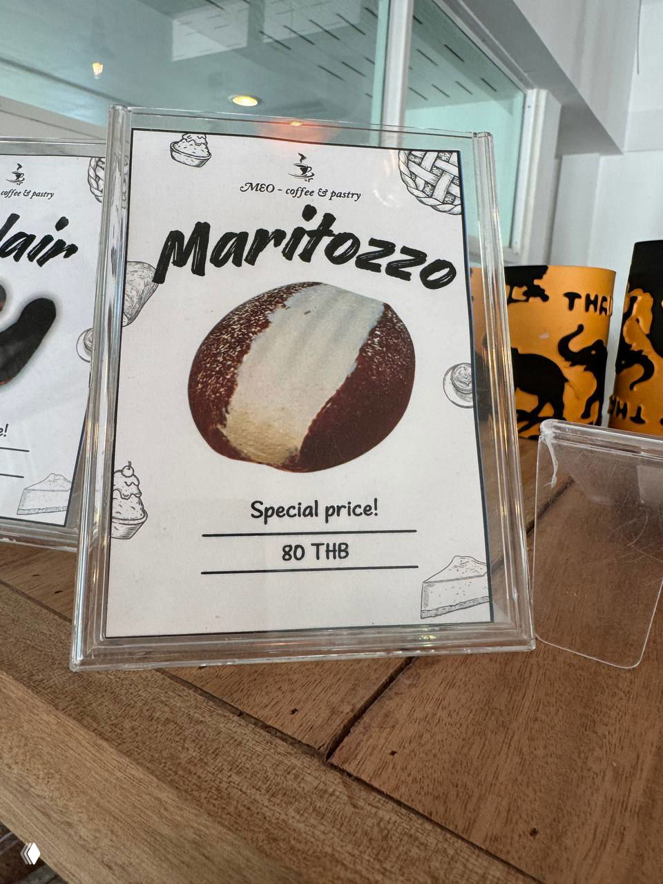 Пластиковая рамка с рекламой булочки Maritozzo и указанием цены 80 THB на деревянном прилавке кофейни, крупный план.