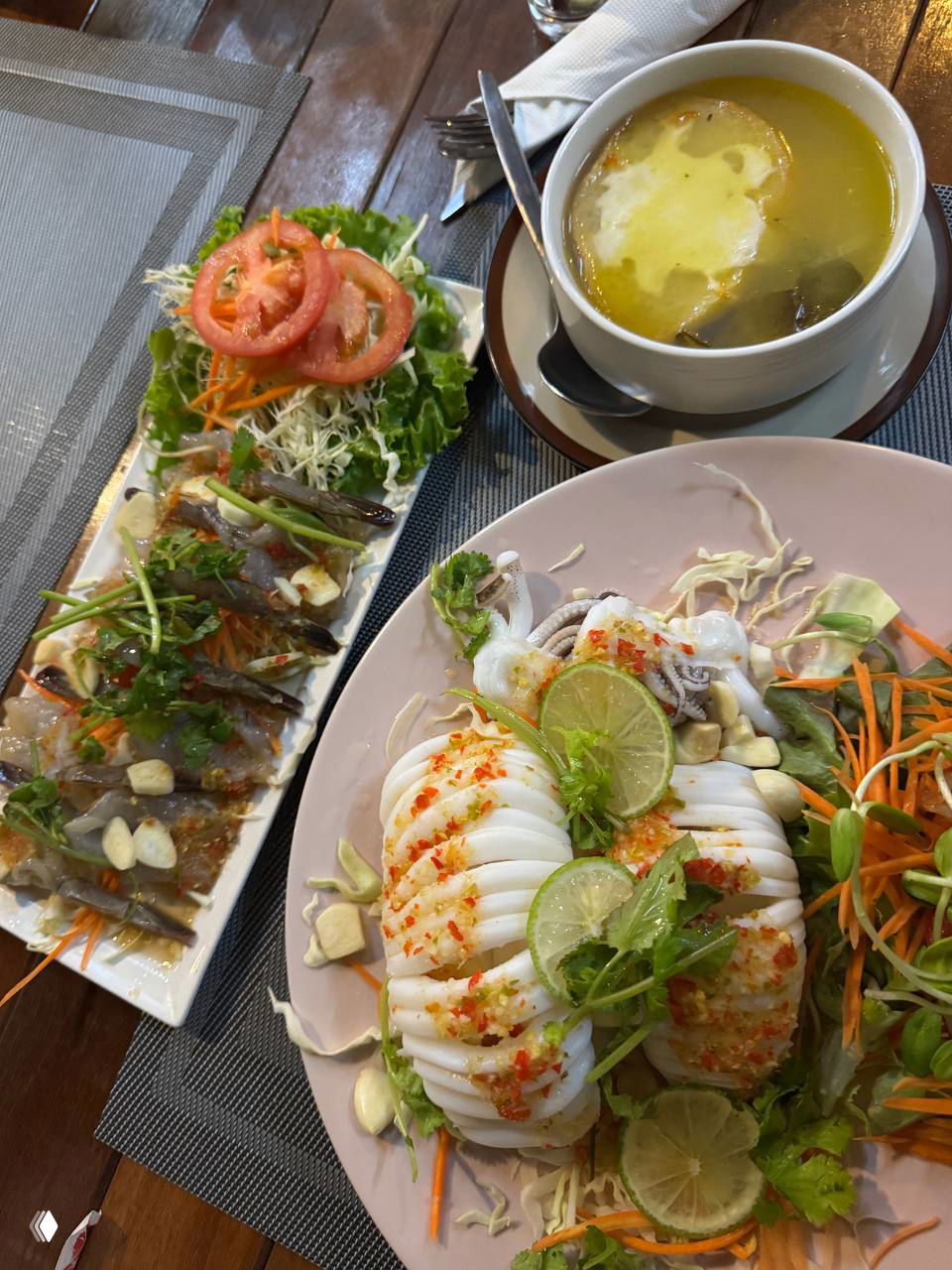 Тарелки с морепродуктами в NOREE: кальмар на пару, сырые креветки и часть лукового супа на деревянном столе с прибором.