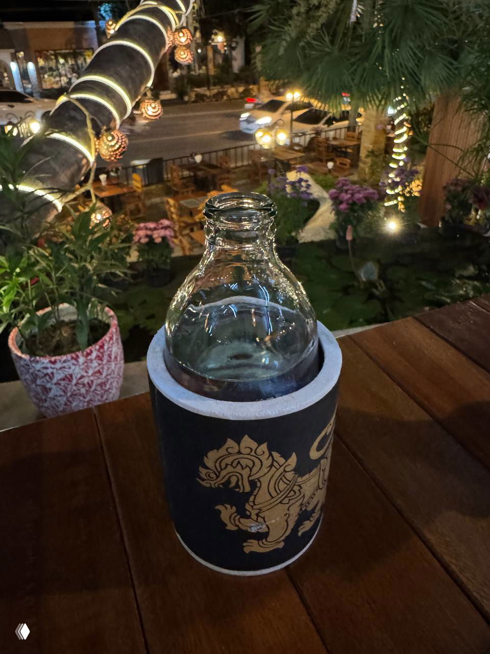 Столик с бутылкой воды в держателе на фоне уличной сцены NOREE: огни дороги, растения и оформленный деревянный интерьер ресторана.