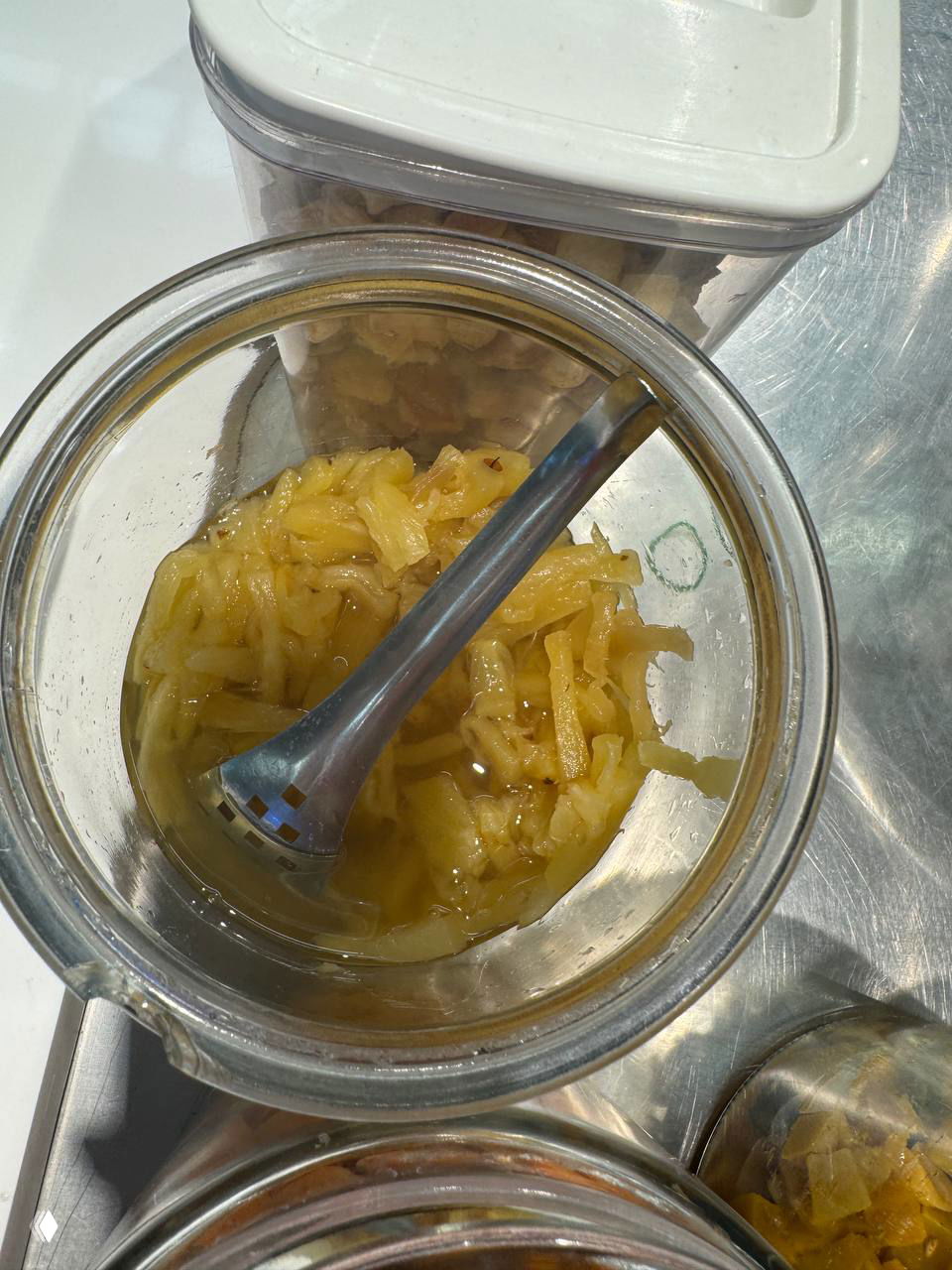 Стеклянная банка с тёртым ананасом в сиропе на прилавке киоска, вид сверху, в кадре ложка для подачи и отражение света