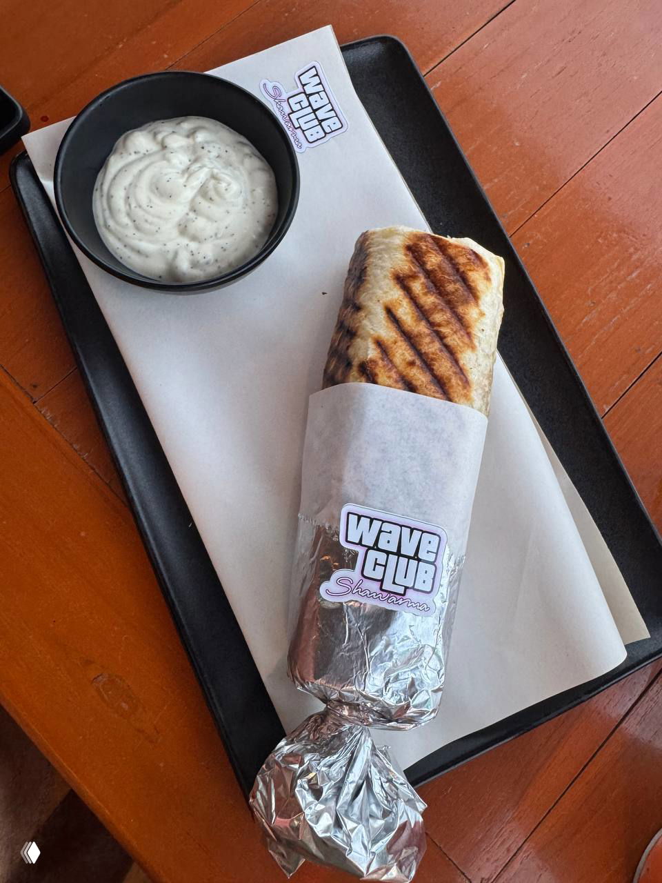 Шаурма Wave Club, аккуратно завернутая в бумагу и фольгу с фирменным стикером Wave Club и мисочка соусa рядом.
