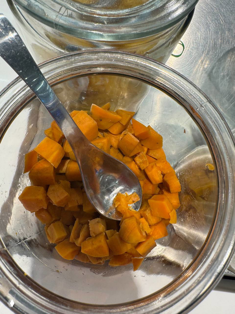 Стеклянная ёмкость с кусочками томлёной тыквы или батата ярко-оранжевого цвета, рядом металлическая ложка для топпингов