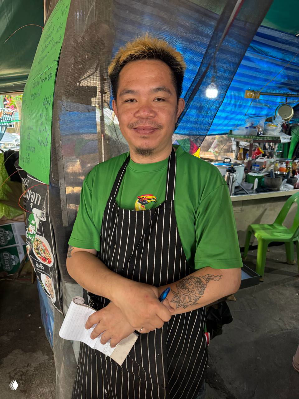 Портрет владельца и повара Tong в черно-белом фартуке у уличного прилавка, улыбается, видна татуировка на предплечье и блокнот в руках.