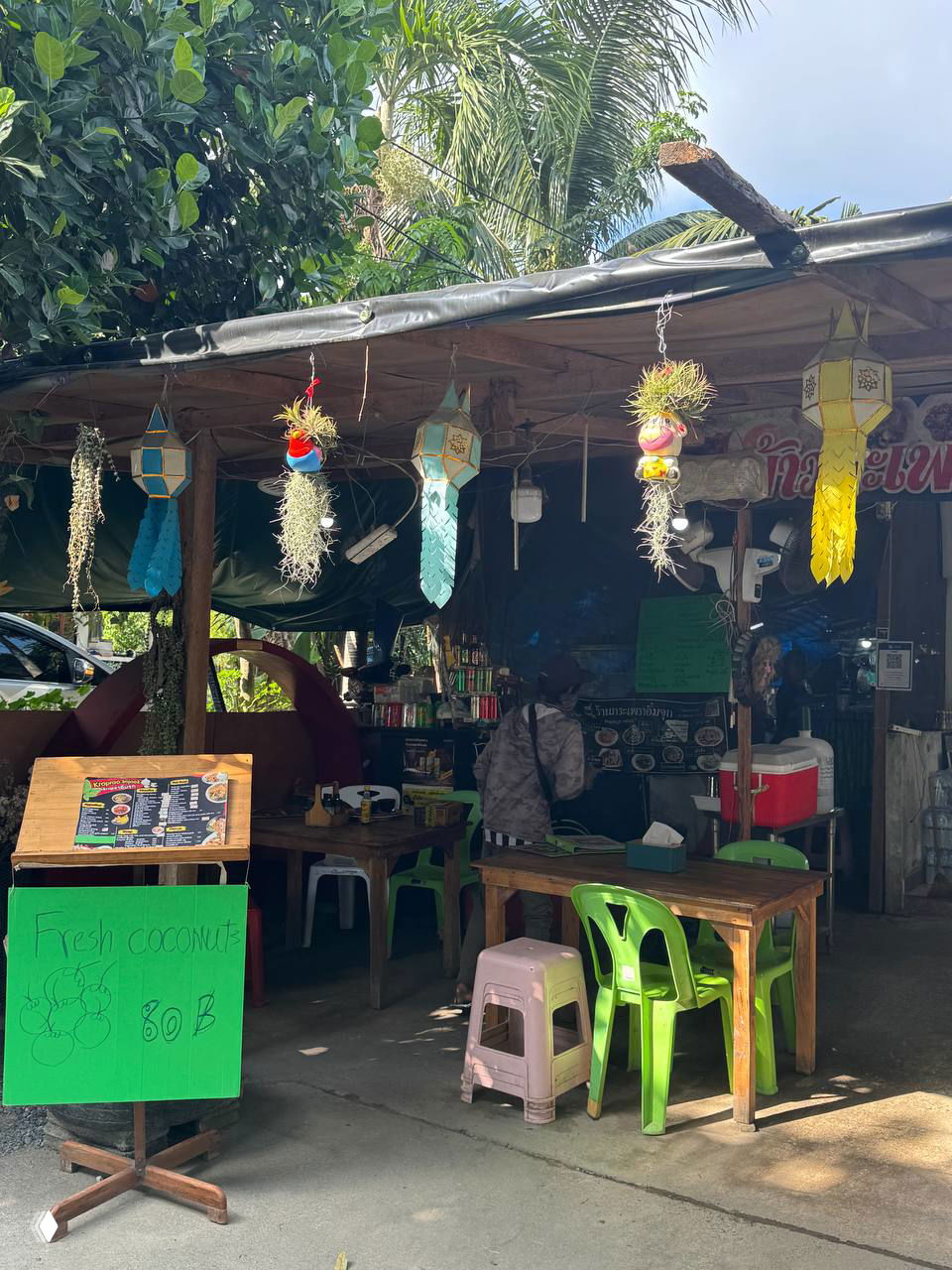 Небольшой уличный ресторан в Ламай: столики с пластиковыми стульями под тентом, вывеска с ценой кокосов и декоративные бумажные фонарики.