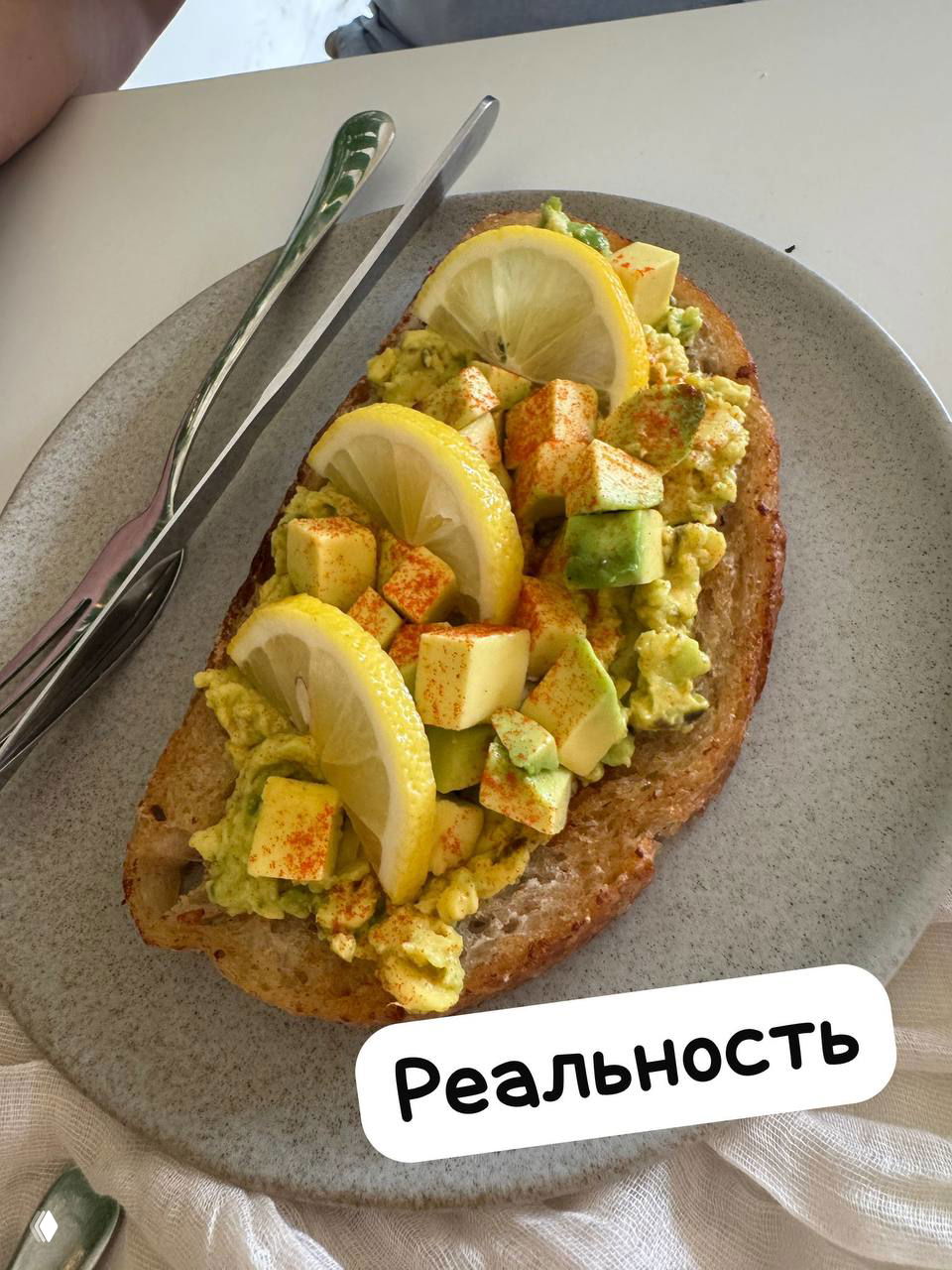 Реальный авокадо-сэндвич на тарелке: рубленое авокадо, кубики авокадо, дольки лимона сверху и специи, подпись «Реальность», фото блюда крупным планом.