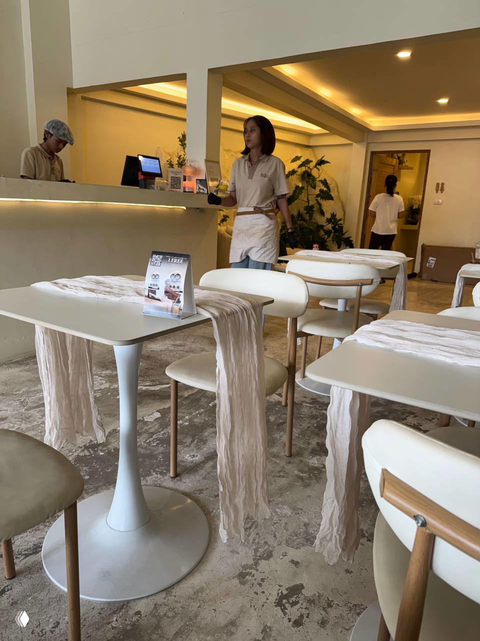 Интерьер кофейни ISLA: светлые белые столы и стулья, стойка и персонал в фирменной форме, минималистичный декор и вид на барную зону.