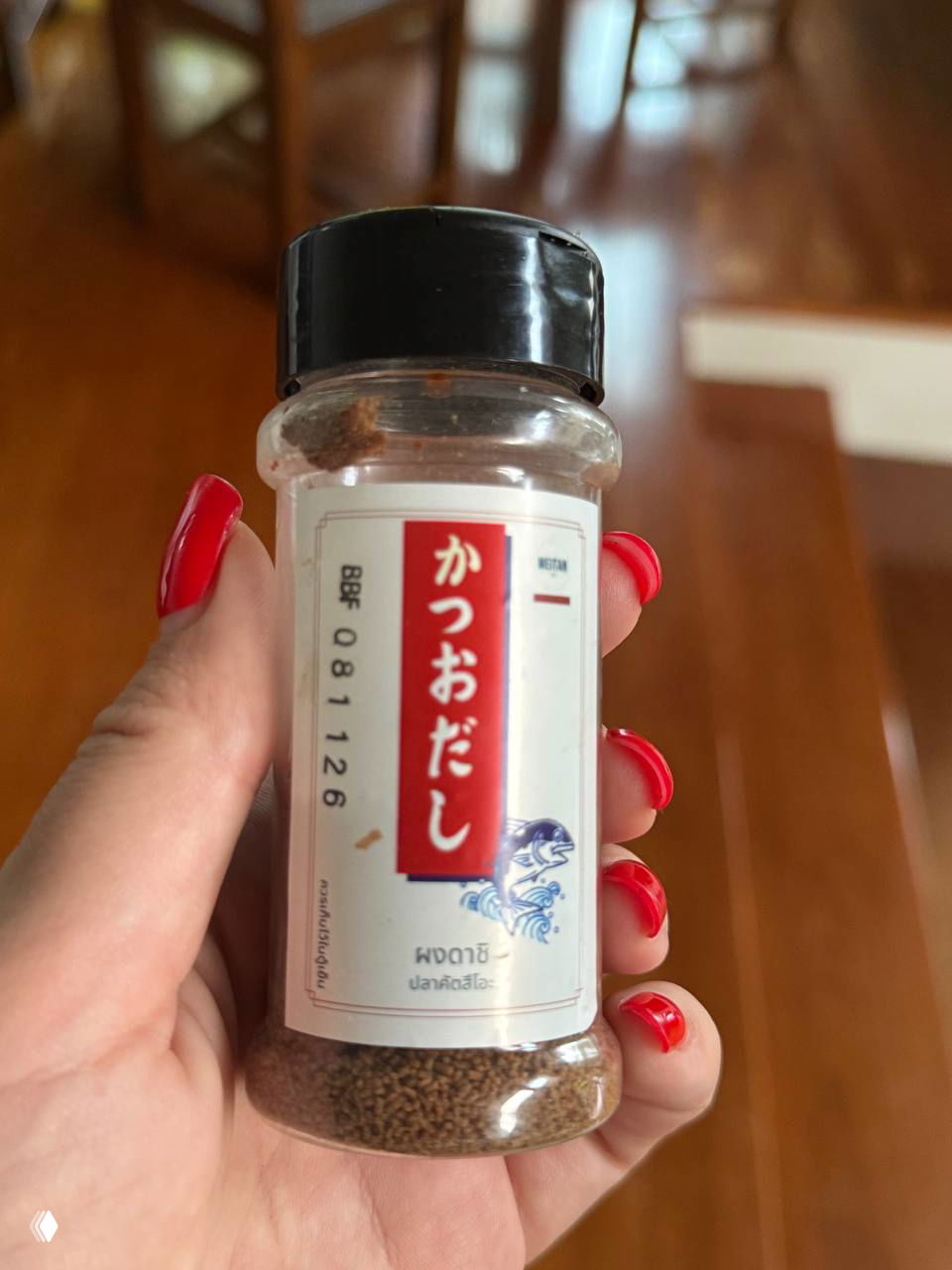 Бутылочка с порошком даси в руке с красным маникюром, видна японская этикетка на белом фоне и деревянный пол в интерьере.