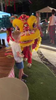 Жёлтый Лев приветствует детей на набережной: праздничная сцена с туристами и семьями, огни и декорации вечернего рынка.