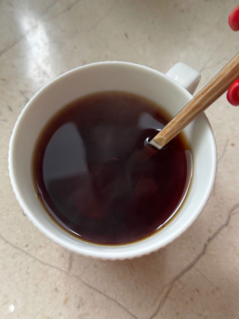 Вид сверху: белая чашка с тёмным заваренным чаем и деревянной ложкой на светлой мраморной поверхности, отражение света на жидкости.