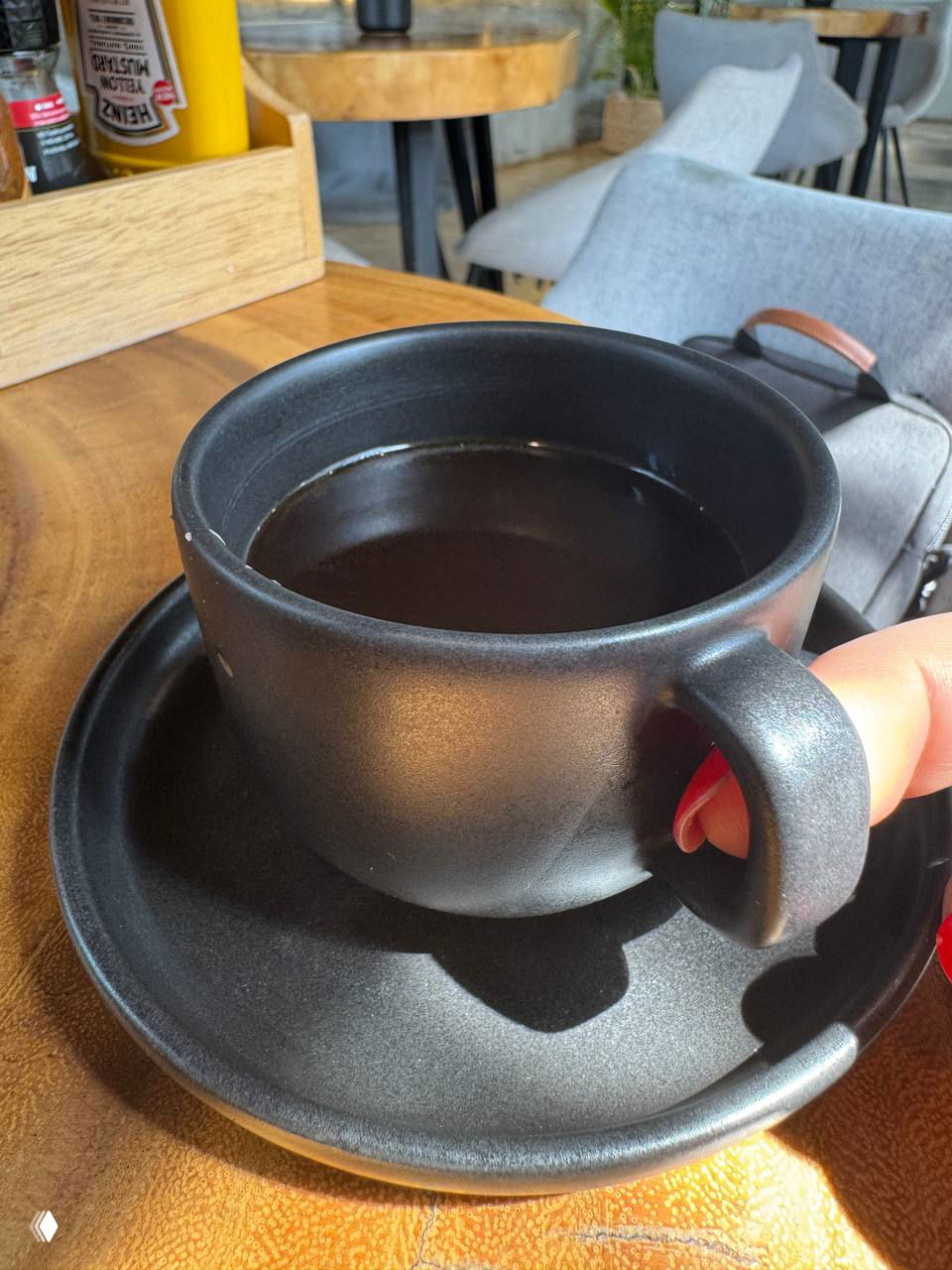 Черная матовая чашка с черным кофе на подходящей подставке-блюдце, рука держит ручку чашки, на фоне деревянного стола и стульев кафе.