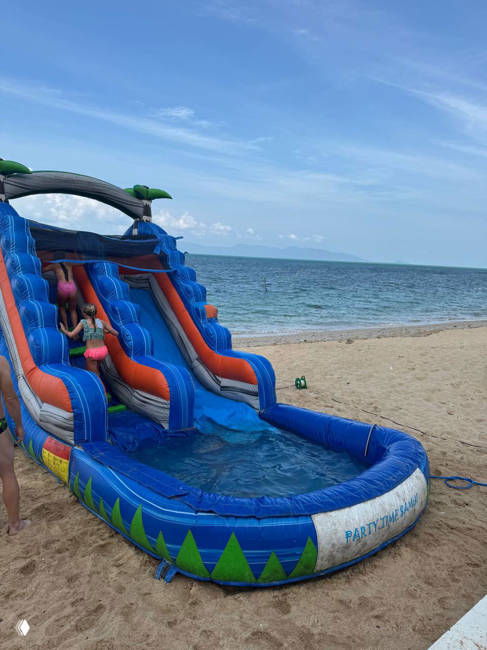 Надувная водная горка на песчаном пляже: дети поднимаются вверх, внизу небольшой бассейн и море на заднем плане — детское развлечение.