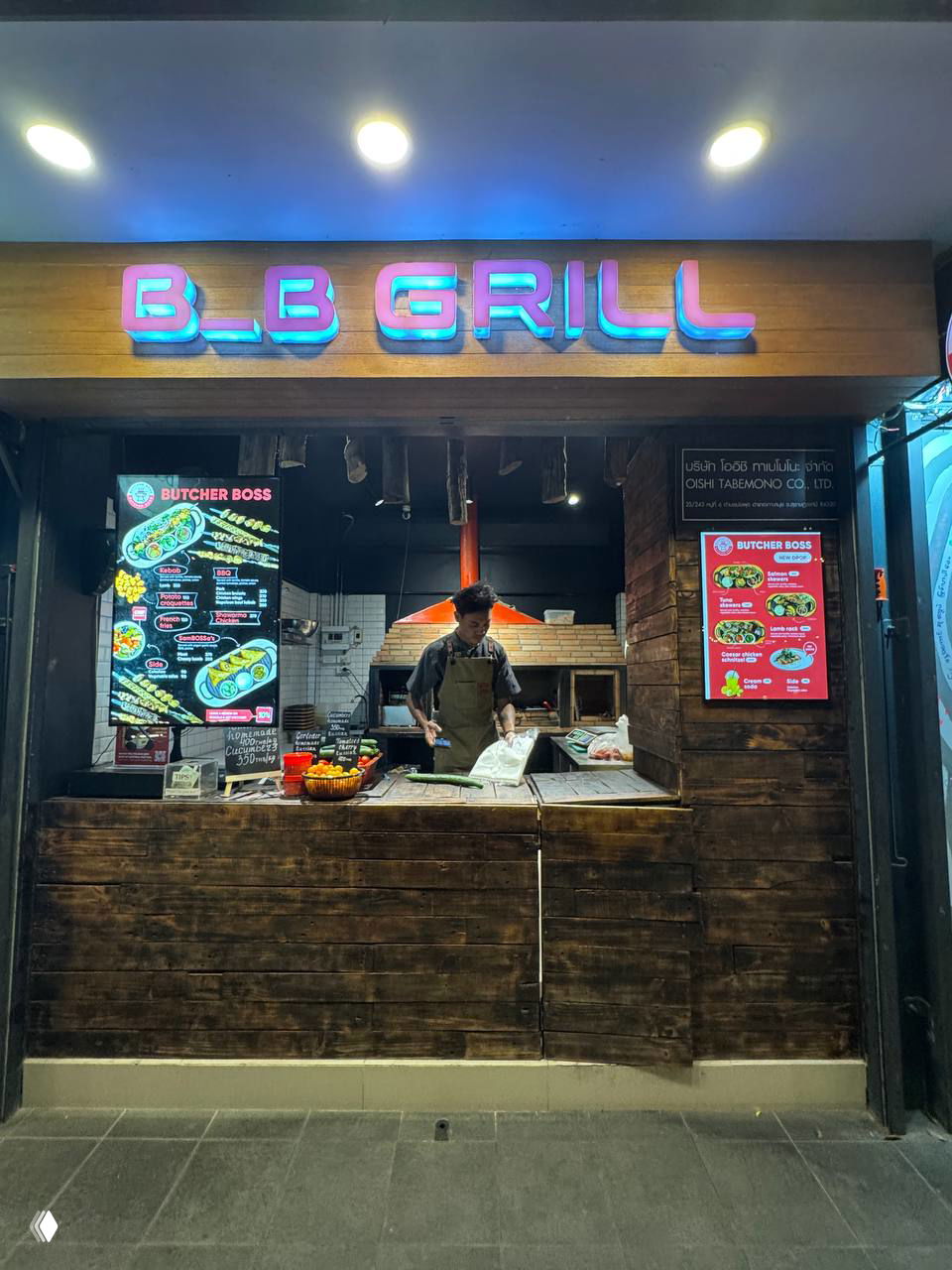 Фасад лавки Butcher Boss / B_B GRILL на рынке: деревянный прилавок, неоновая вывеска и повар, готовящий блюда у печи вечером.