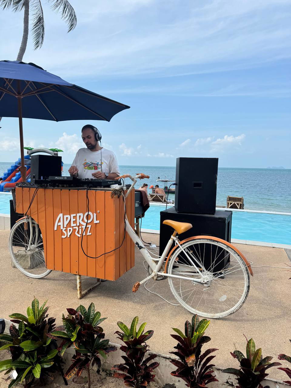 Диджей у бассейна за стойкой с надписью Aperol Spritz; рядом велосипед-стойка и акустическая аппаратура на фоне моря — музыкальная зона.