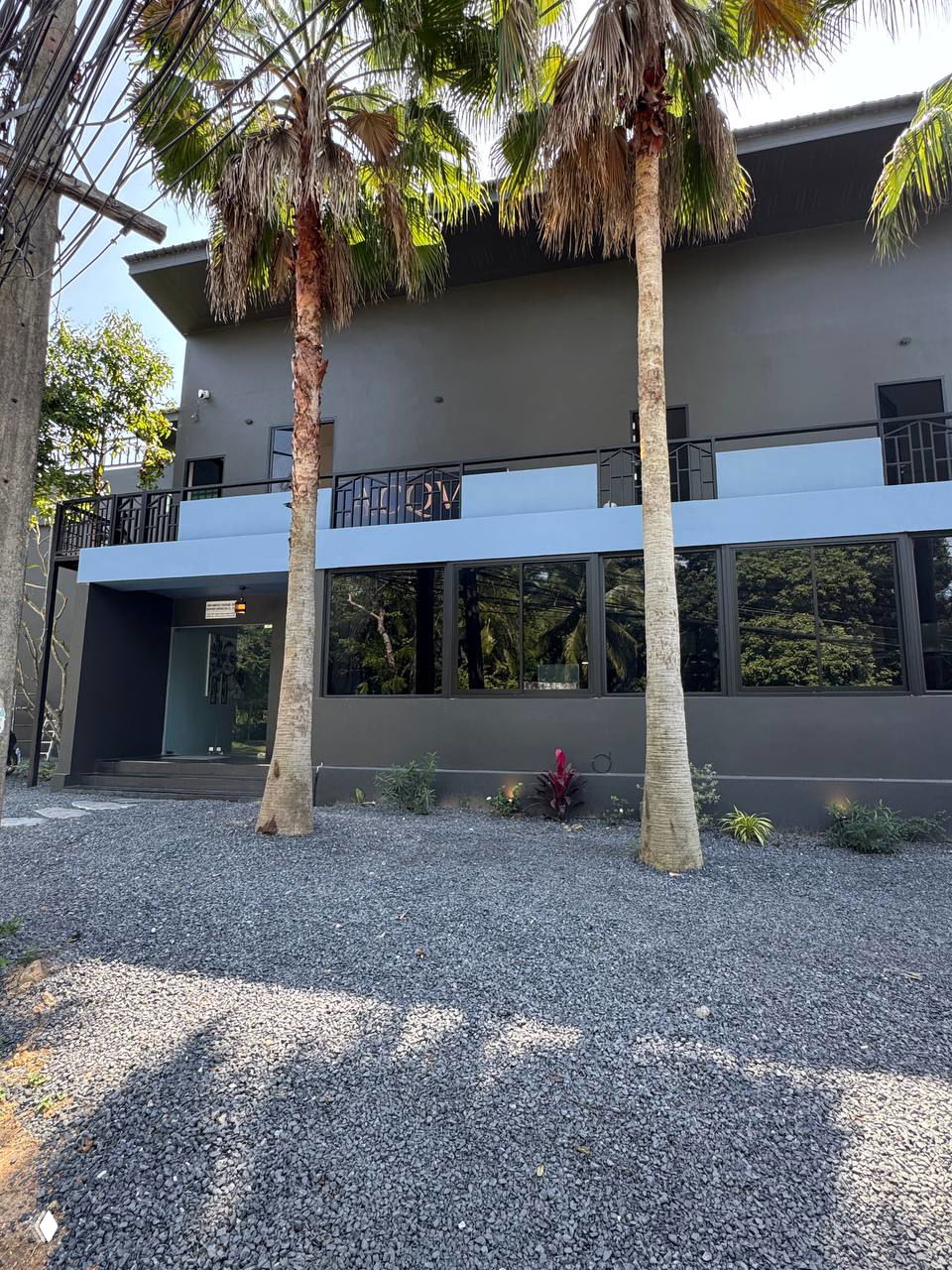 Фасад и стоянка перед зданием ресторана ACQUA в Бопуте: двухэтажное серое здание с большими окнами и пальмами спереди.